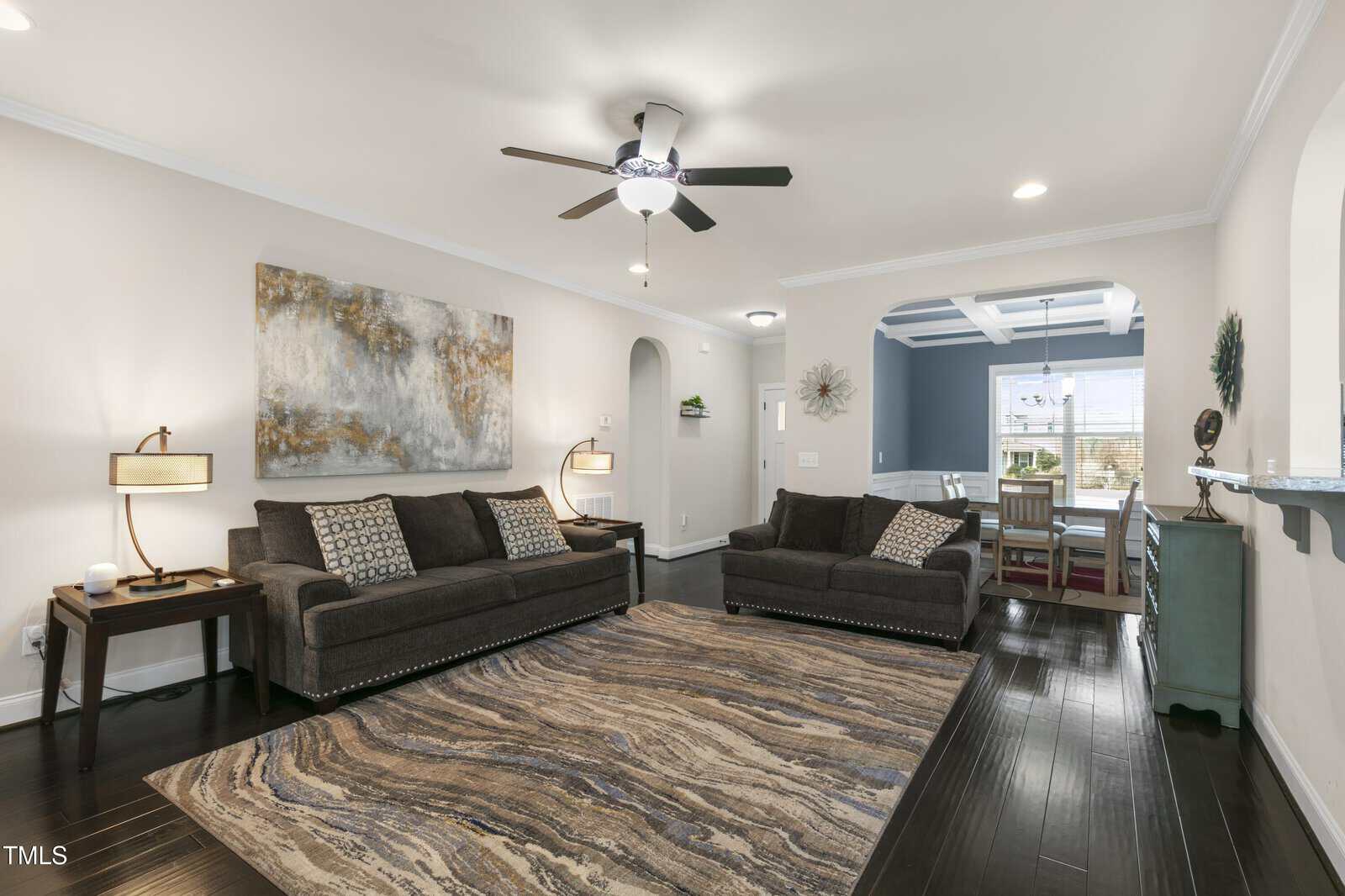 A living room with dark wood flooring, a large abstract painting, two gray sofas with patterned cushions, and a ceiling fan. A patterned rug sits in the center, and an archway leads to a dining area with a table and chairs.