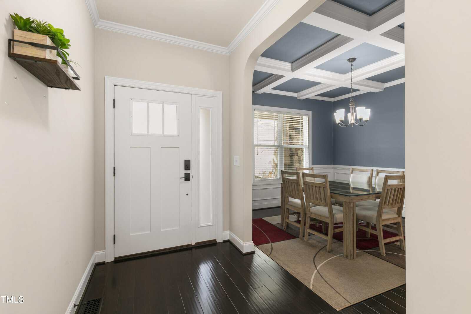 Hallway leading to a dining room with a coffered ceiling and chandelier. The dining area features a wooden table with chairs on a beige and red rug. Walls are painted in a light and dark color contrast. A potted plant sits on a wall shelf.