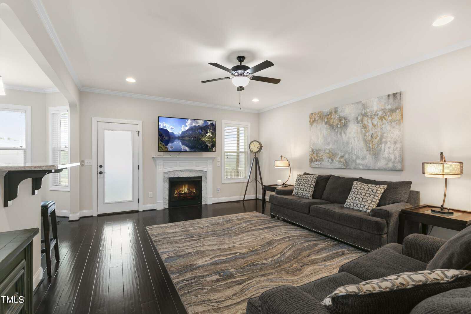 A living room with a dark wood floor features a gray sofa set, a large abstract painting, and a rug. There's a fireplace beneath a wall-mounted TV and a ceiling fan above. Two lamps and a standing clock complete the decor.