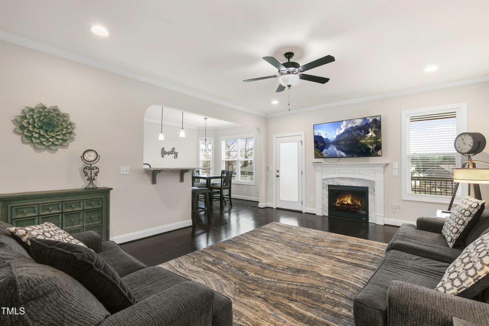 A living room with gray sofas, a patterned rug, and a white fireplace with a TV above it. A ceiling fan is on, and there's a dining area visible through an archway. Walls are beige, and windows provide natural light. Decorative items adorn the space.