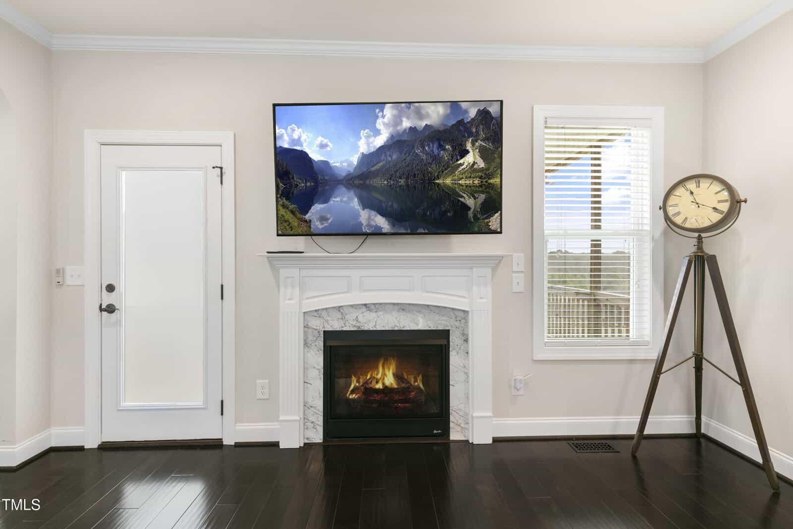 Living room with a wall-mounted TV above a fireplace displaying a mountain lake scene. To the right, a large clock on a tripod stand. A door with frosted glass and a window with blinds are on the left. The floor is dark hardwood.