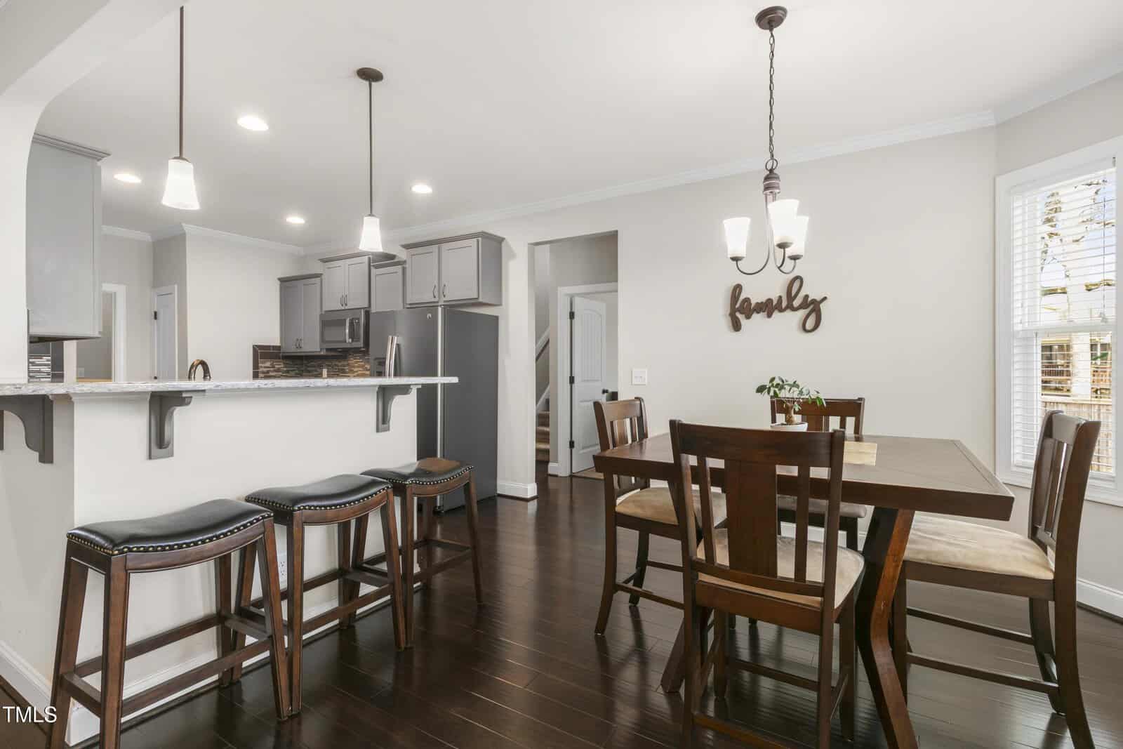 A modern kitchen and dining area with dark hardwood floors. The kitchen features an island with four bar stools and gray cabinets. The dining area has a wooden table and six chairs, with a chandelier above. A "family" sign adorns the wall.