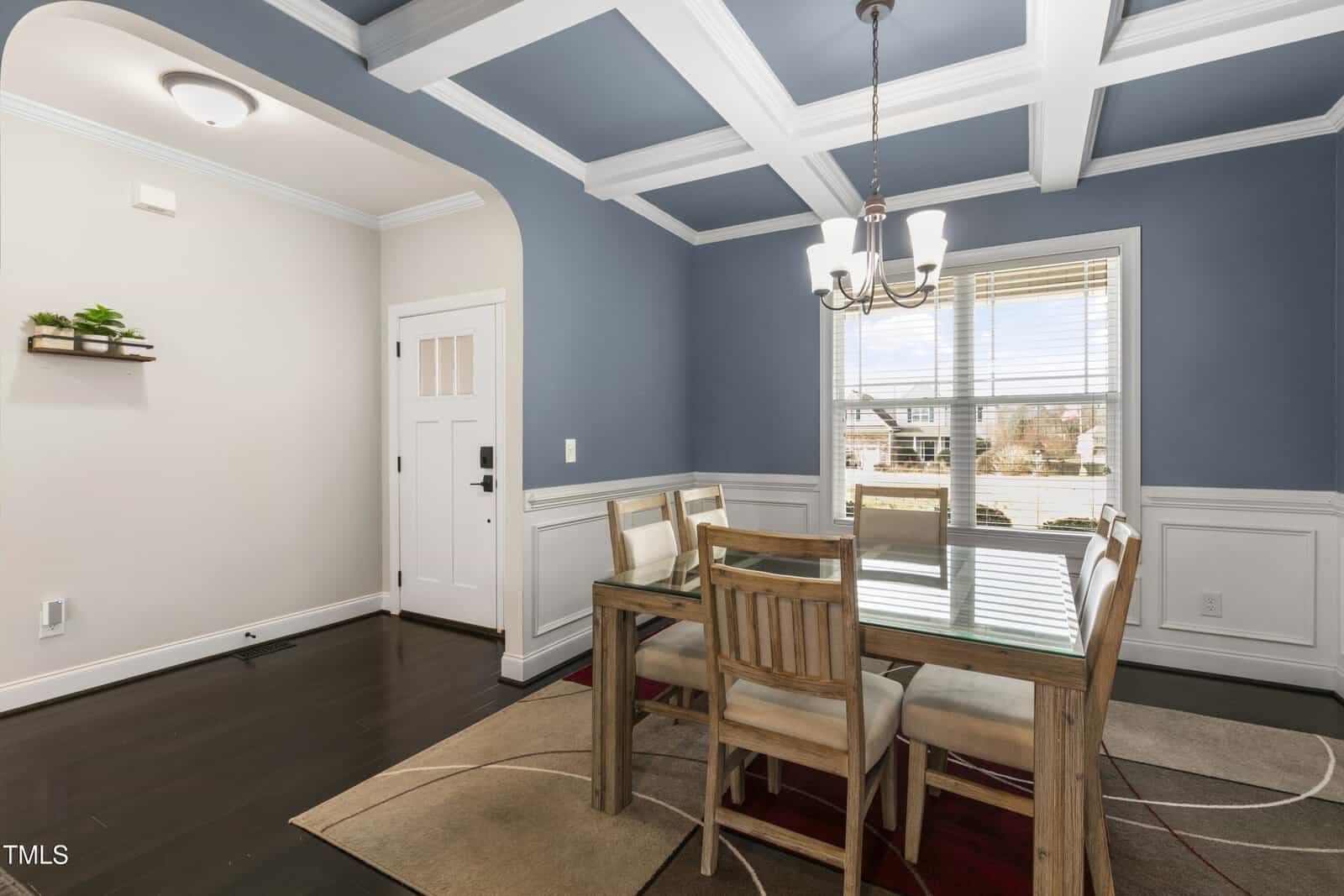 A dining room with dark wood flooring features a wooden table and four chairs on a patterned rug. The walls are painted blue and white, with a coffered ceiling. A window with blinds and a light fixture above the table complete the setting.