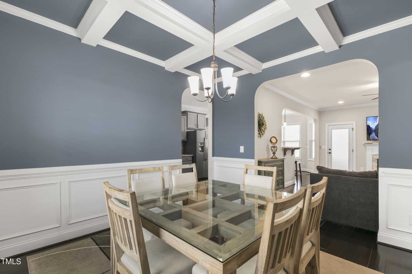 Dining area with a glass-top wooden table and six chairs on a rug. The room features blue walls, white wainscoting, and a coffered ceiling with a chandelier. An open doorway leads to a kitchen, visible with white cabinetry.