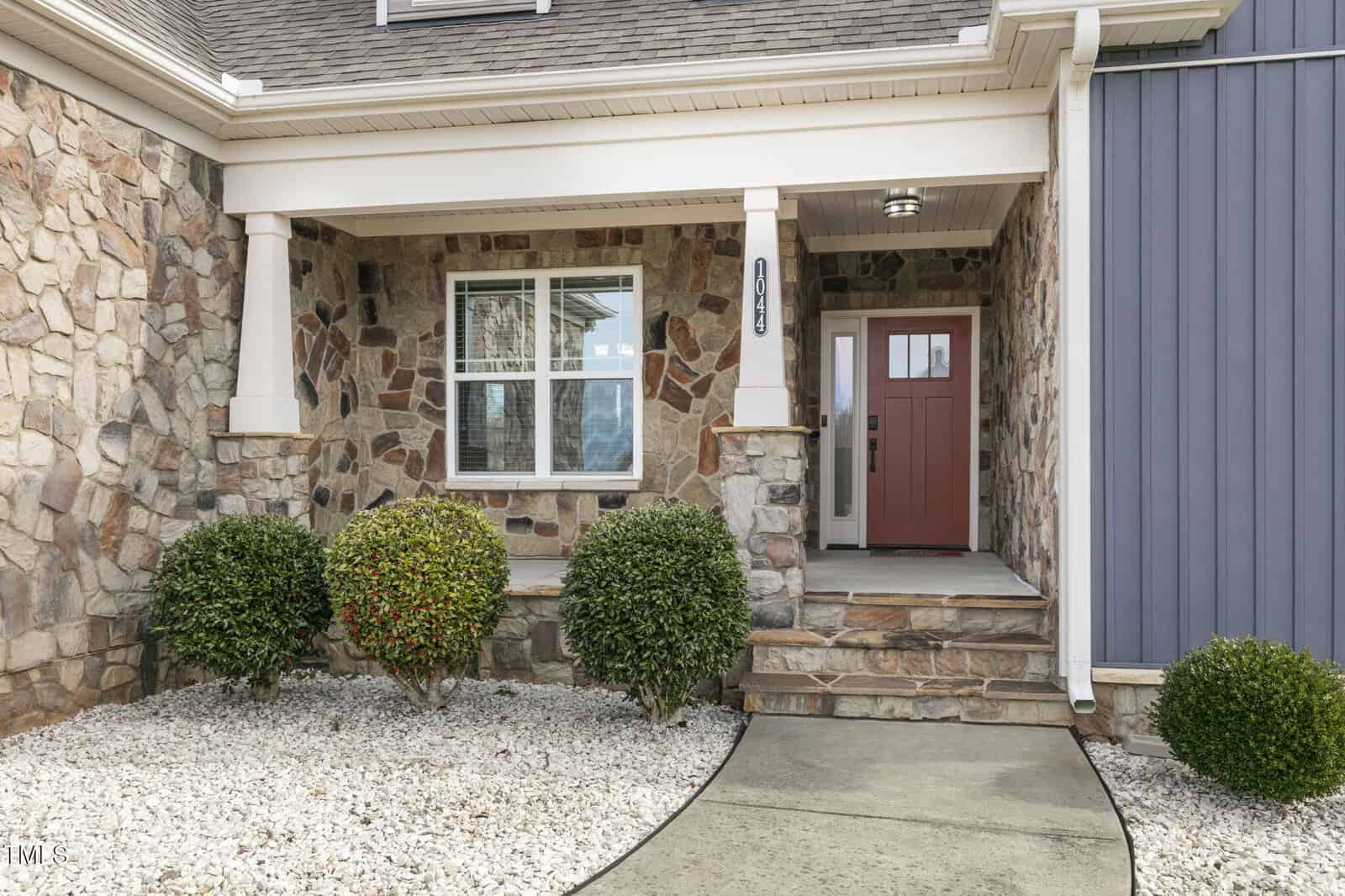 Front entrance of a house with stone facade. A red door is centered between a window and a white column. Two bushes sit on the pebble-covered ground, bordered by a concrete walkway. Blue siding is visible on the right side.