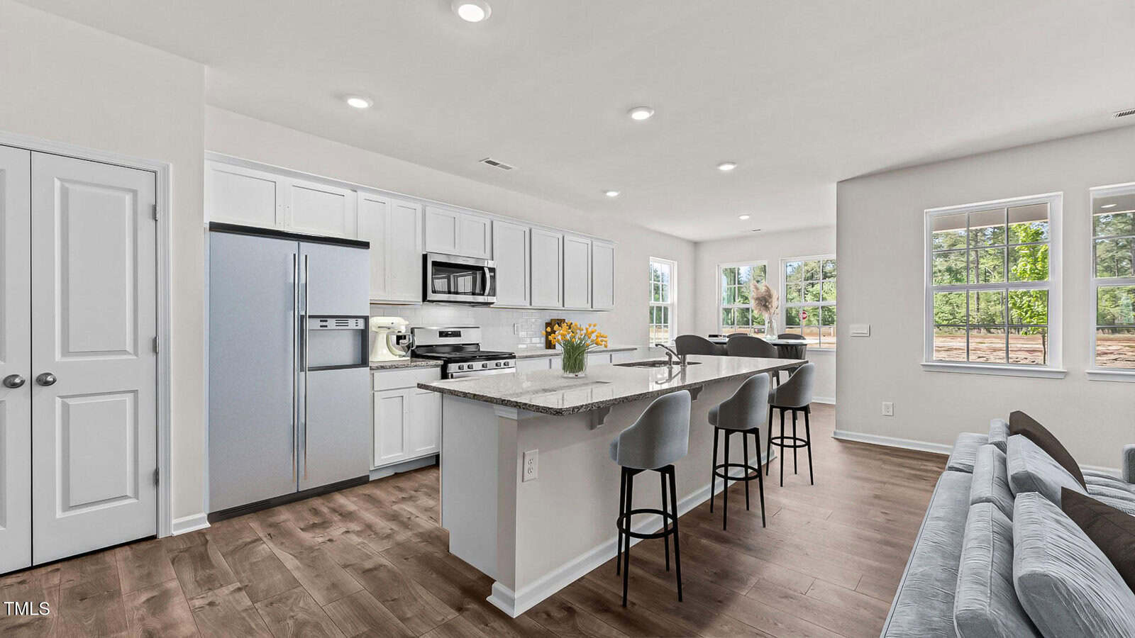 A modern kitchen with white cabinets, stainless steel appliances, a large island with barstools, and a vase of flowers. Adjacent to the kitchen is a living area with a sofa and large windows letting in natural light.