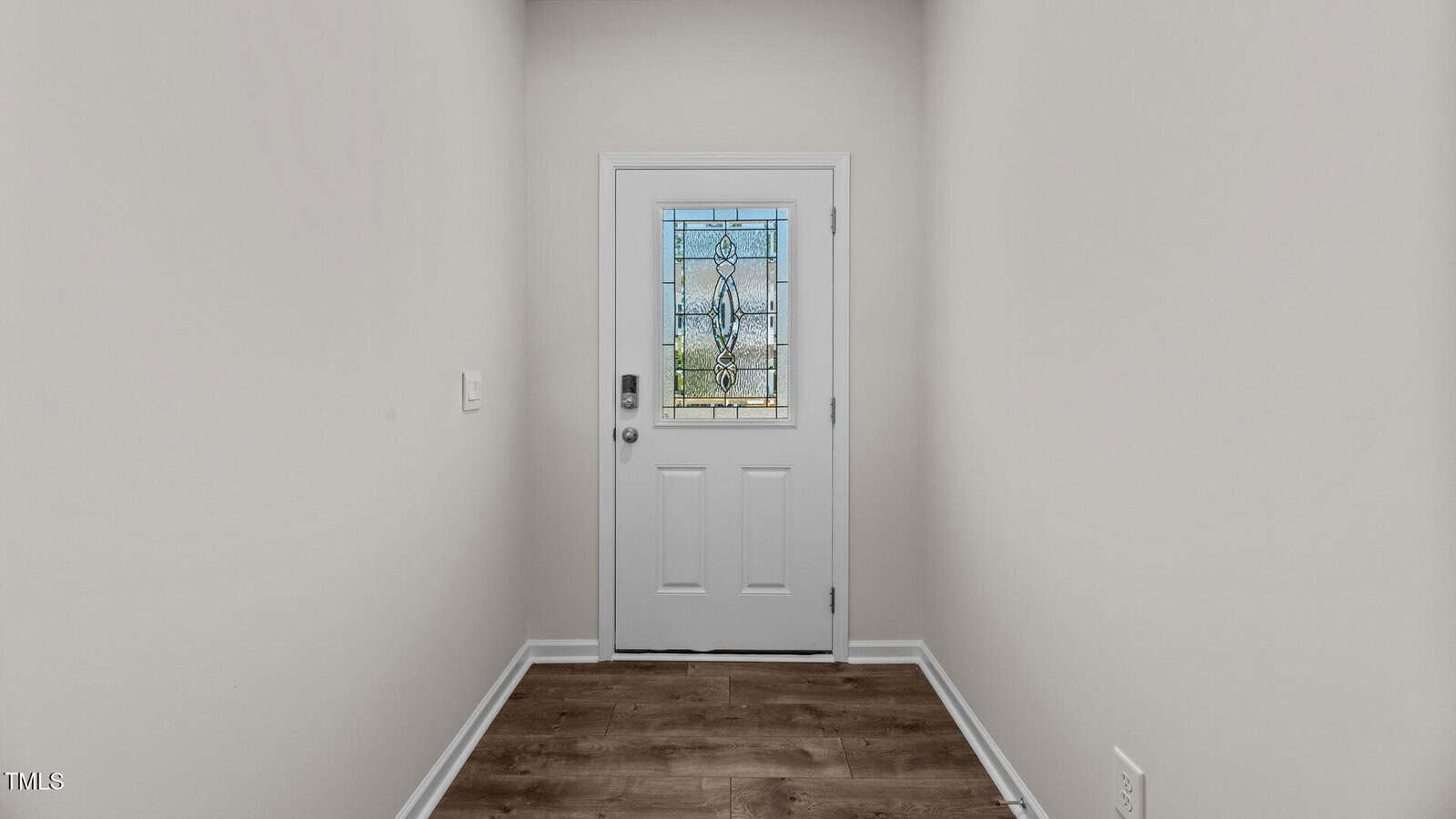 A narrow entryway with light-colored walls, wood-style flooring, and a white front door featuring a decorative glass window. There is a light switch on the left wall and an electrical outlet on the right.