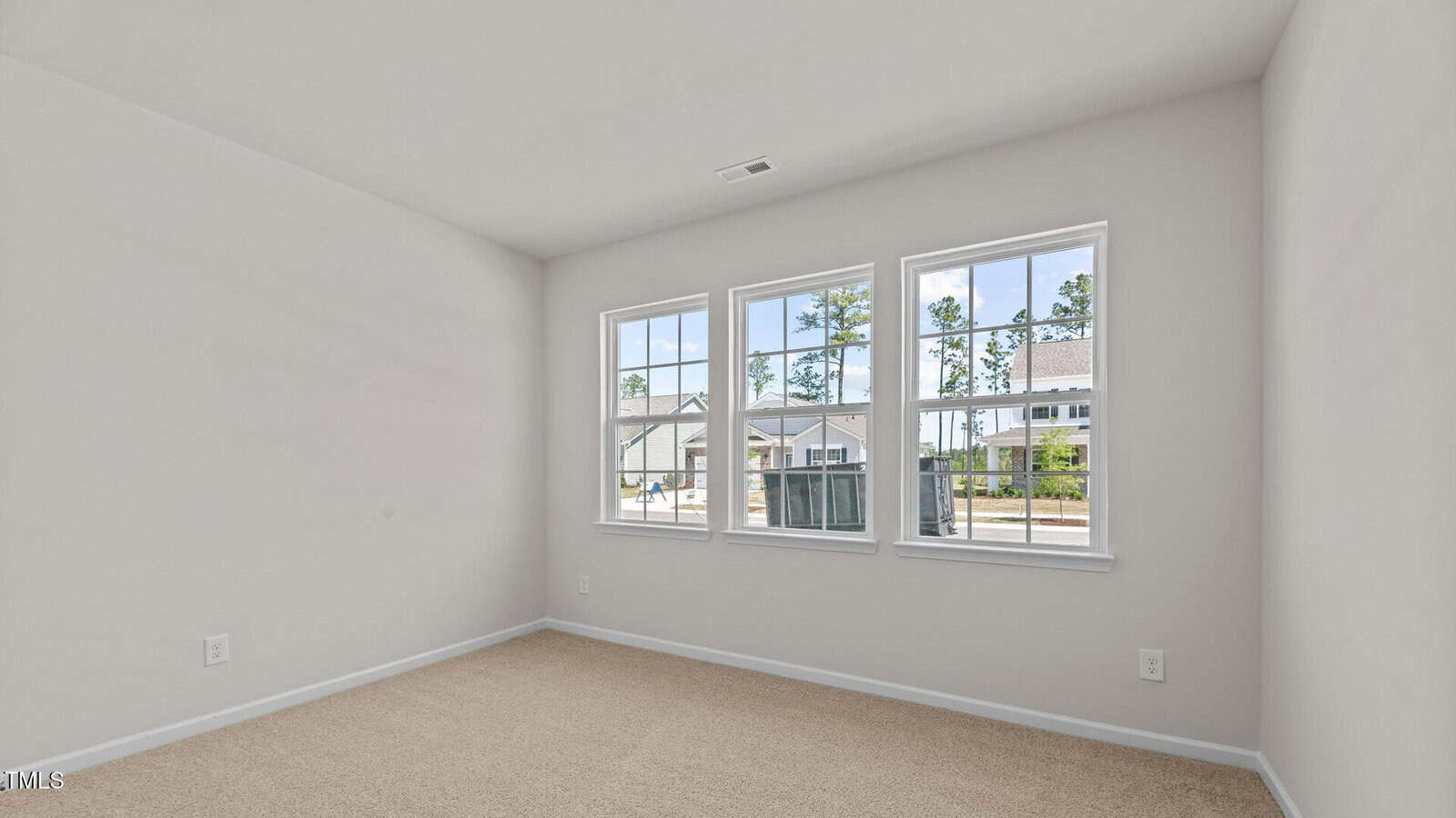 Empty room with light beige carpet, off-white walls, and three large windows on one wall. Outside the windows, neighboring houses, trees, and a street are visible. The ceiling is plain with a single vent.