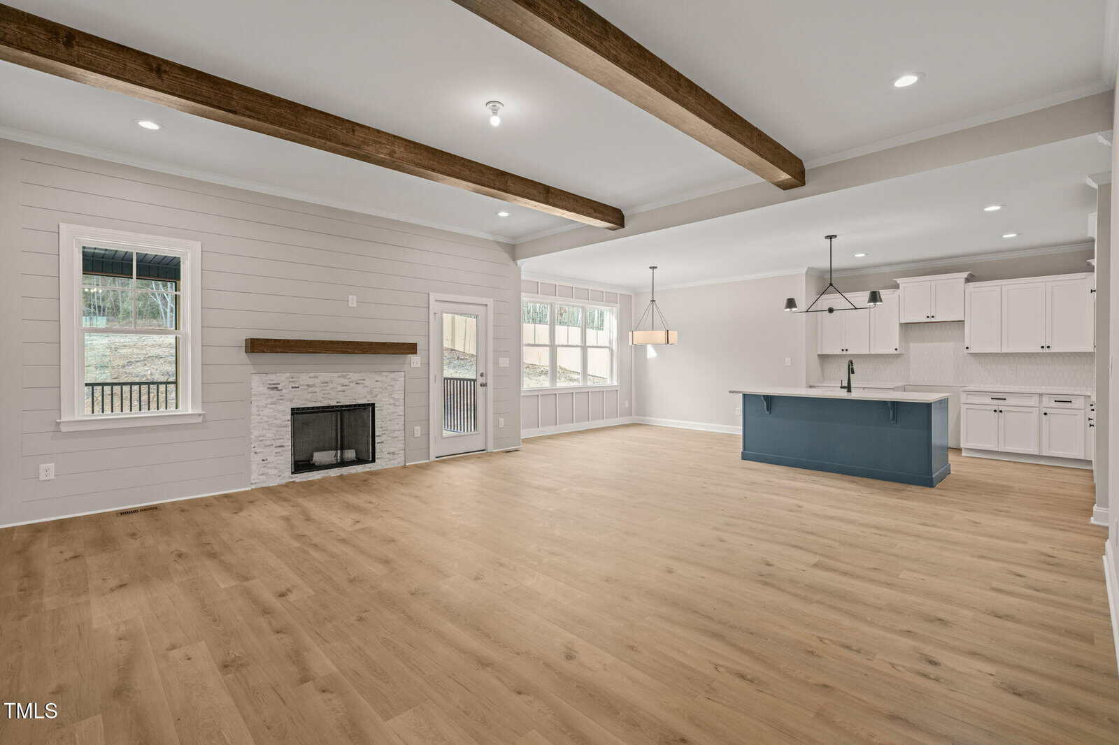 Spacious open-concept living area with light wood floors, white walls, exposed ceiling beams, a fireplace with a white mantel, and a modern kitchen featuring white cabinets and a blue island with a sink.