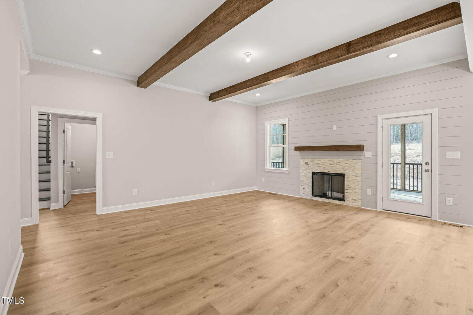 Spacious living room with light wood flooring, white shiplap walls, exposed wooden ceiling beams, a fireplace with a mantel, window, glass door, and an open doorway leading to a stairwell.