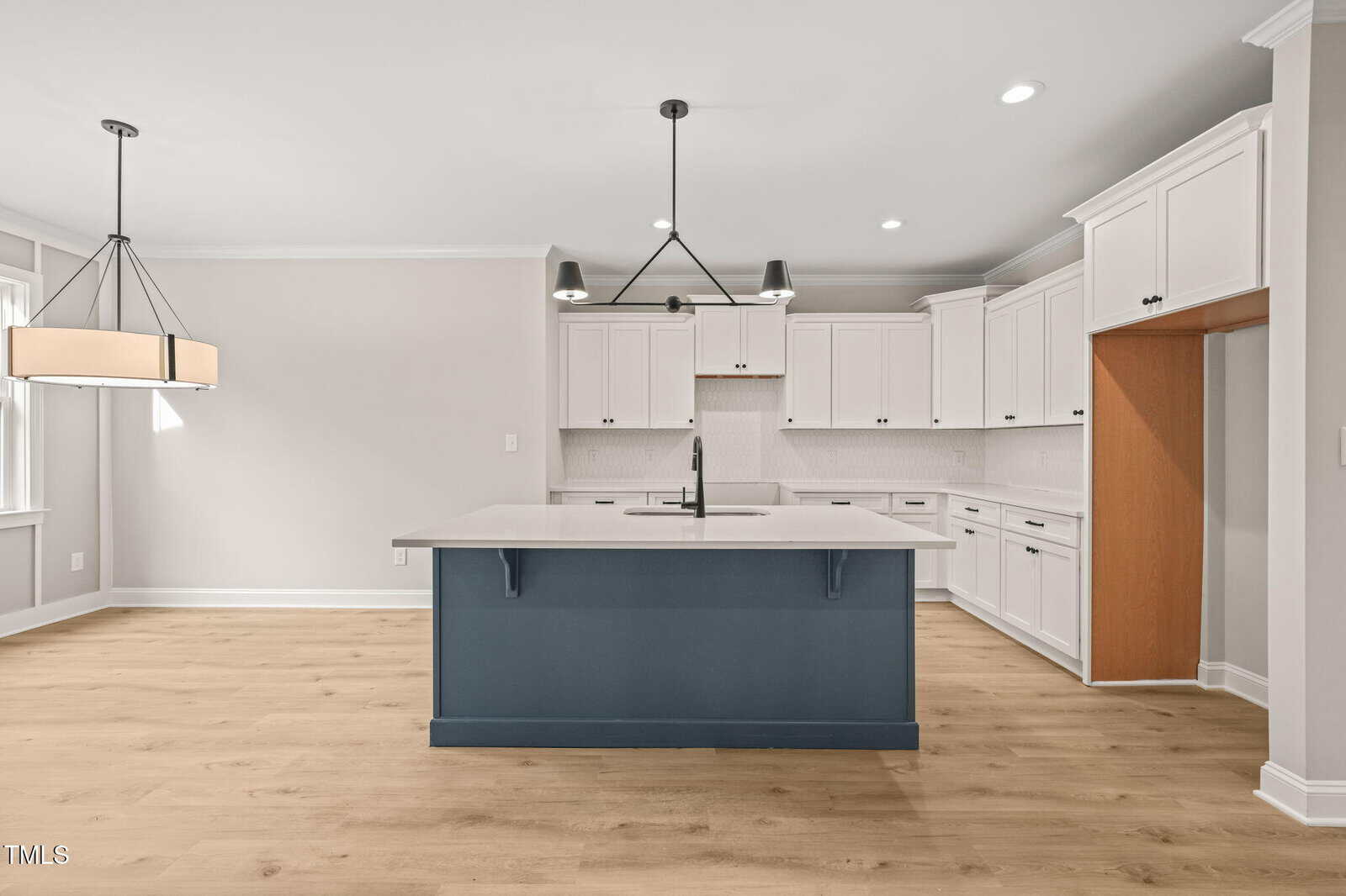 Modern kitchen with white cabinets, a blue island with a sink, light wood flooring, black pendant lights above the island, and a large empty space for a refrigerator on the right. The room is brightly lit with natural and recessed lighting.