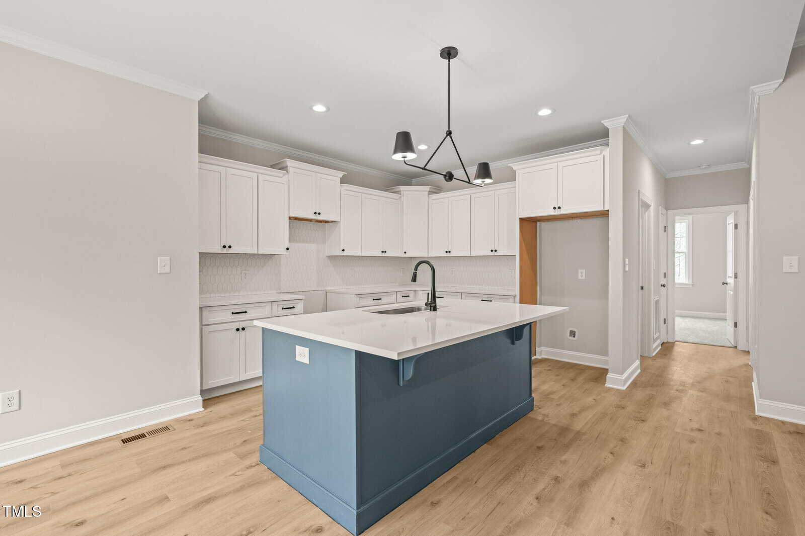 Modern kitchen with white cabinets, a blue island with a white countertop, black faucet, light wood flooring, white walls, and recessed lighting. Black chandelier hangs above the island. Doorways lead to other rooms.