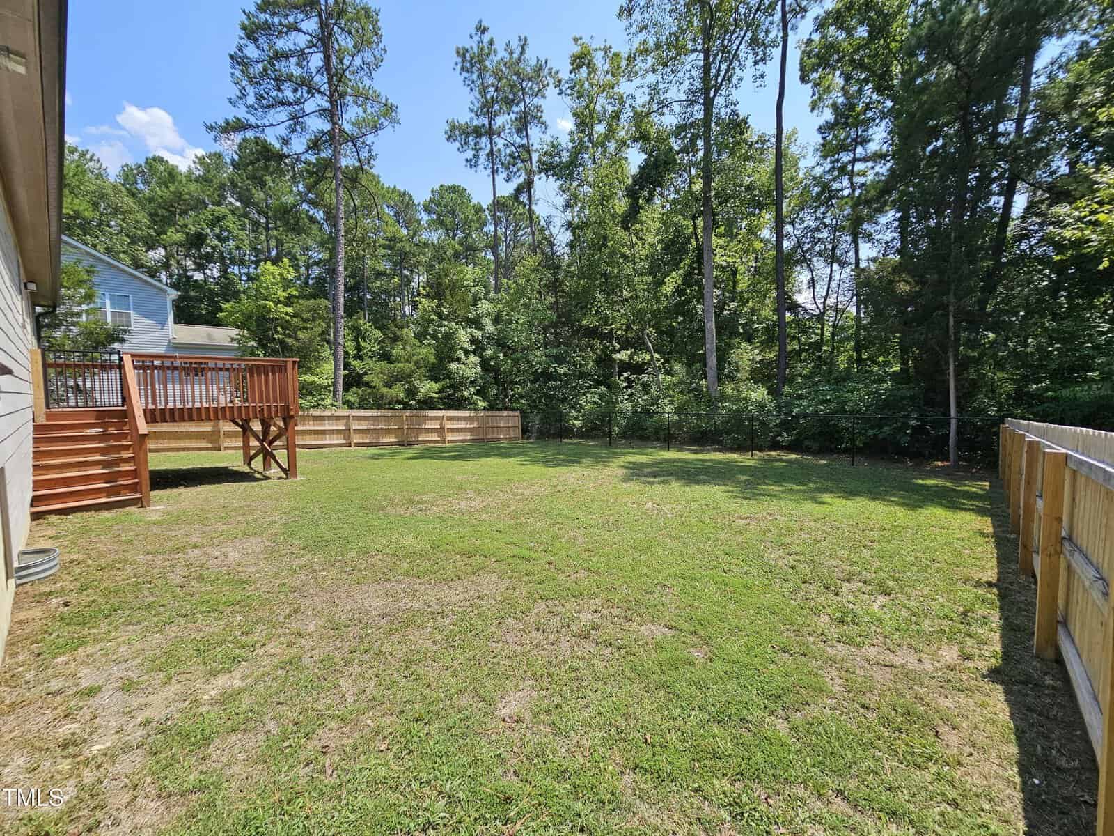A spacious backyard with a partially grass-covered lawn, surrounded by a wooden fence. On the left, there is a house with a wooden deck. Tall trees line the back of the yard beneath a clear blue sky.