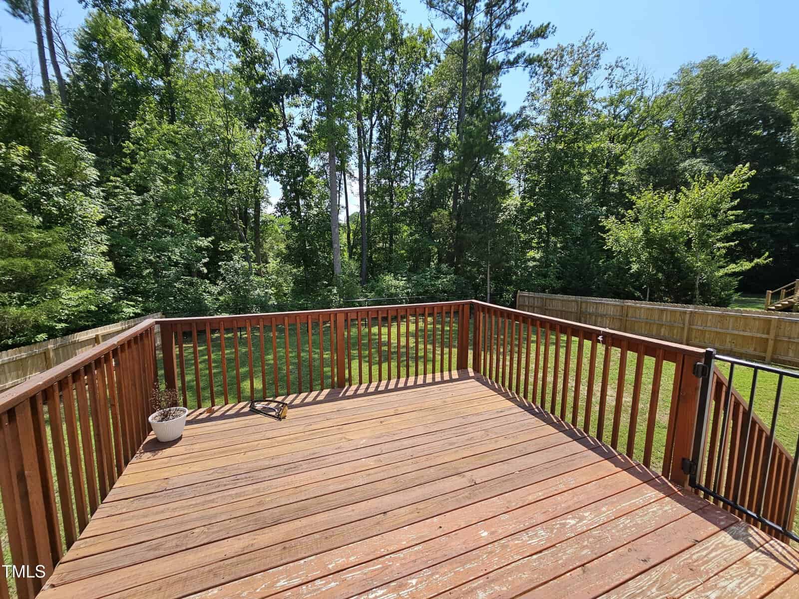 A wooden deck extends into a backyard, bordered by a wooden fence. The yard is grassy and surrounded by a dense line of trees. A single small potted plant is placed on the deck. The sky is clear and blue.