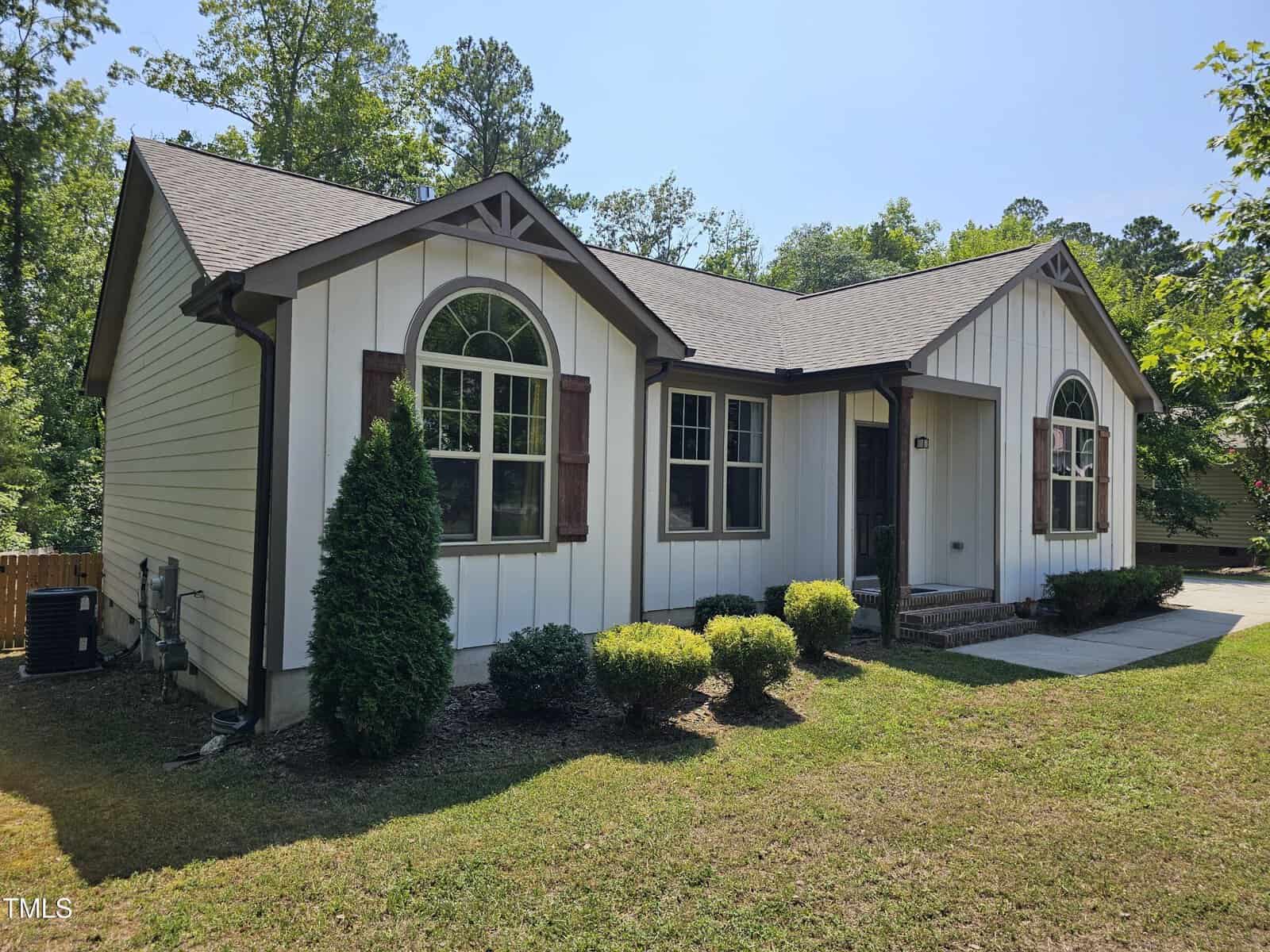 A small, single-story house features white siding with brown trim and accents. It has three arched windows at the front and a gabled roof. The house is surrounded by a grassy lawn with some shrubs and trees in the background.