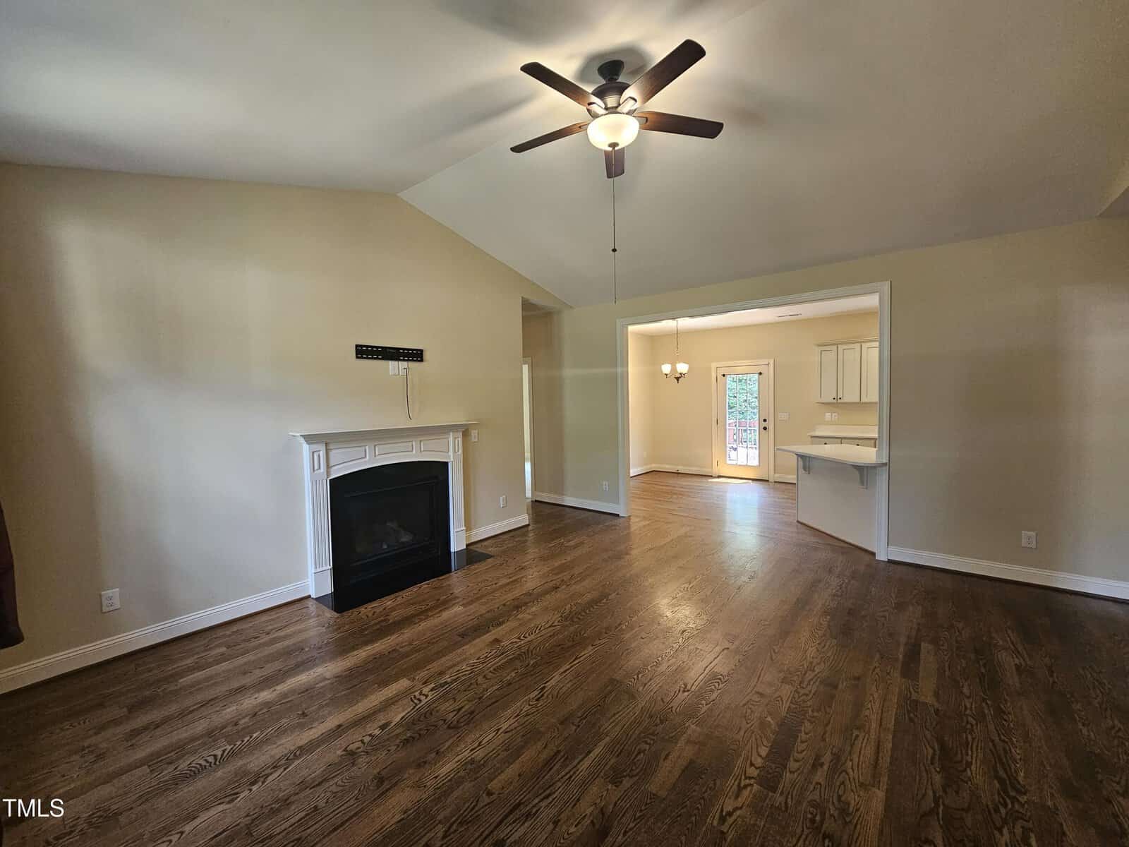 A spacious living room with hardwood floors and a ceiling fan. A fireplace is on one wall. An open doorway leads to a dining area with a chandelier and a door to the outside. The walls are painted a light beige color.
