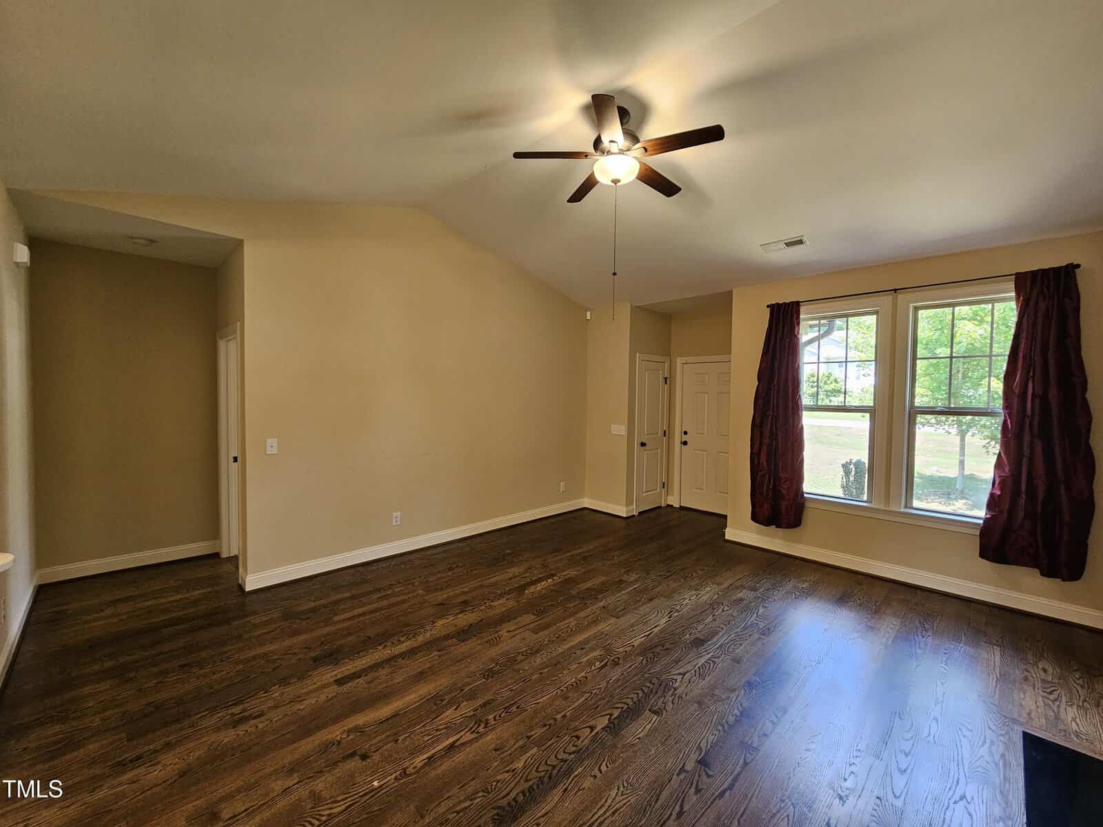 A room with hardwood flooring, beige walls, and a ceiling fan with lights. Large window with dark red curtains on the right. A closed door is also visible under the window. The ceiling is sloped, enhancing the room's spaciousness.