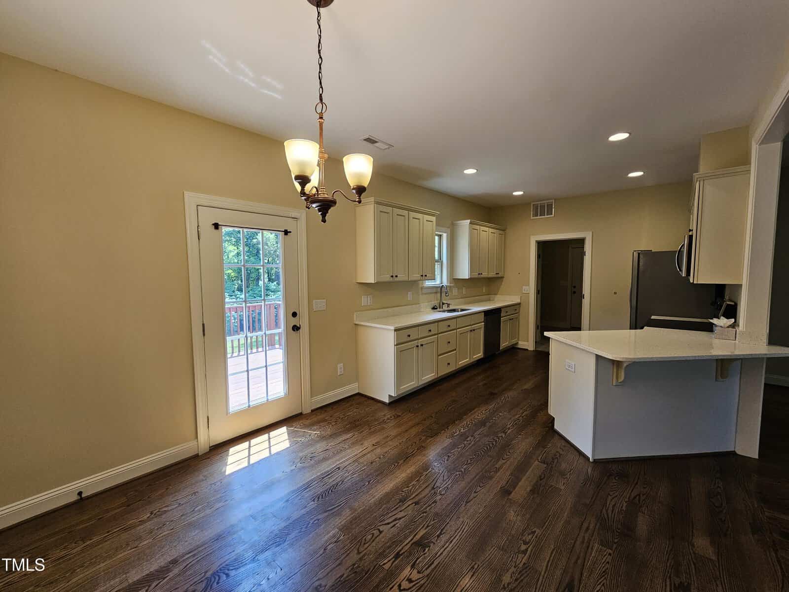 A kitchen with hardwood floors, light tan walls, and white cabinets. There's a central island with a white countertop, a stainless steel refrigerator, and a dining area with a door leading to a deck. Ceiling lights illuminate the space.
