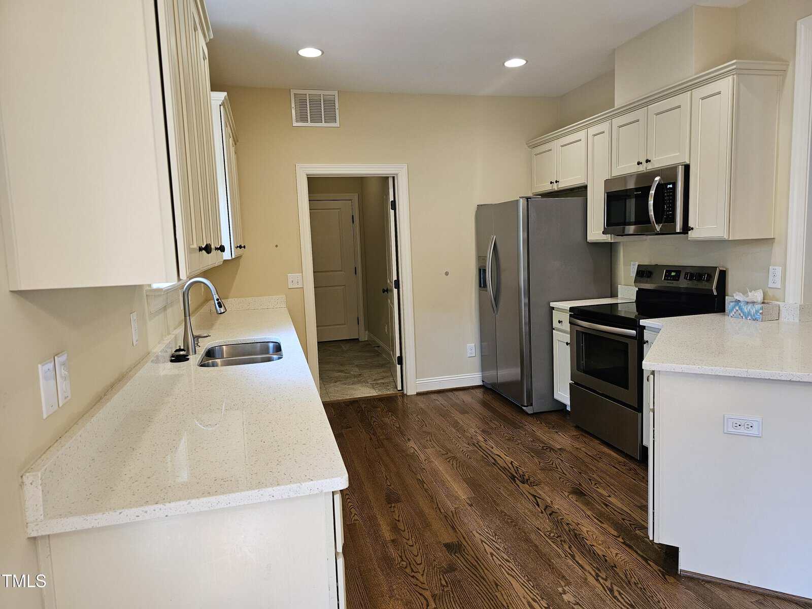 A kitchen with white cabinets, stainless steel appliances including a refrigerator, oven, and microwave. The countertops are a light stone material, and the floor is hardwood. A sink is set into one of the countertops. A doorway leads to another room.