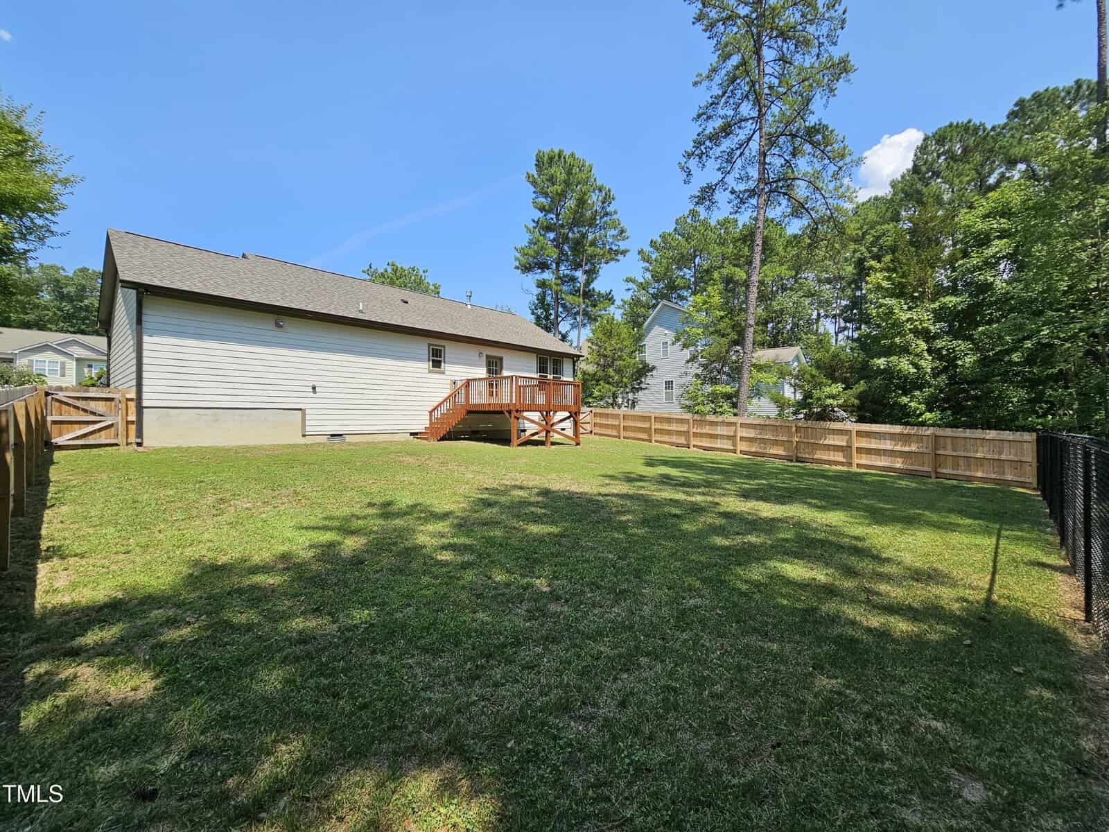 A spacious backyard with a grassy lawn is bordered by a wooden fence. In the background, there is a light-colored house with a sloped roof and a wooden deck. Tall trees are visible around the perimeter, under a clear blue sky.