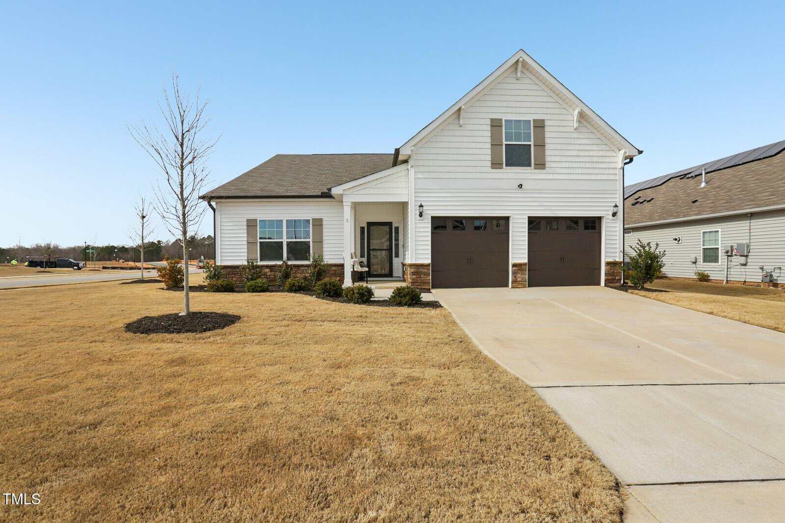 A single-story white house with a double garage, stone accents, and a covered front porch sits on a large, dry, yellow lawn with minimal landscaping under a clear blue sky.