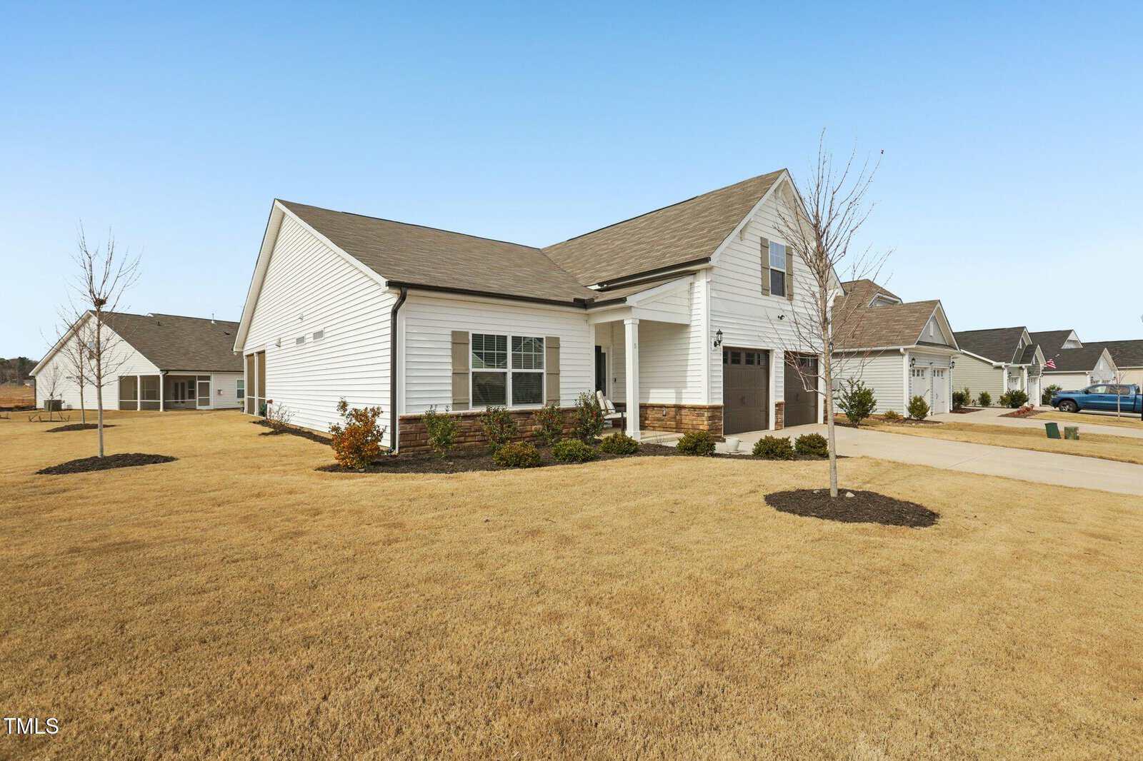 Single-story white house with a gabled roof, attached garage, and stone accents on the exterior. The lawn is brown and has a few small trees and shrubs. The house is in a suburban neighborhood on a clear day.