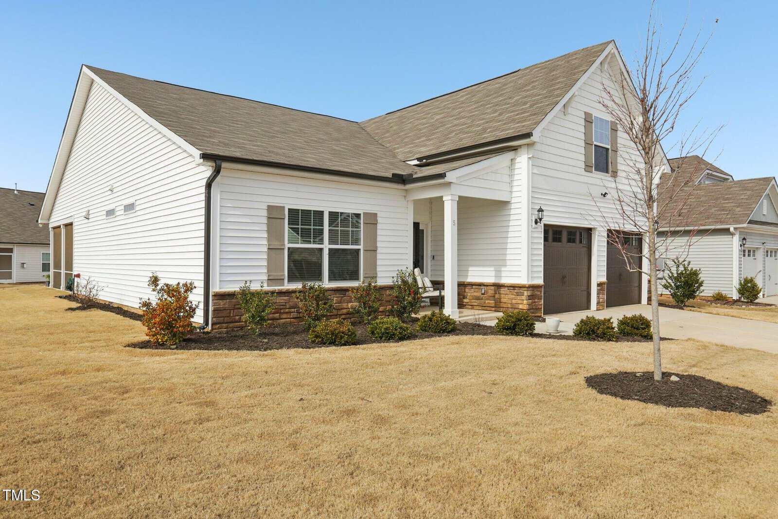 Single-story white house with gray shingles, stone accents, and an attached garage. The front yard has dry grass, a few small shrubs, and a leafless tree. The sky is clear and blue.