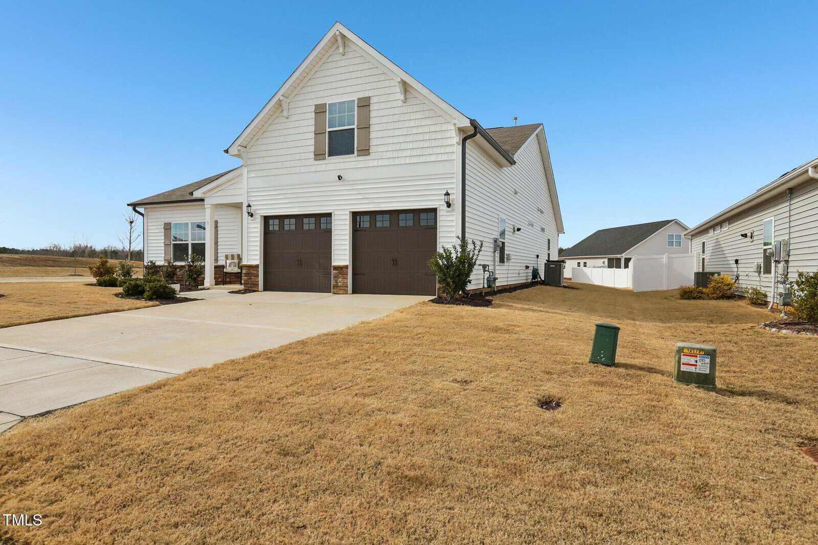 A white single-family house with a two-car garage, brown doors, and a large, dry grass lawn under a clear blue sky. The driveway extends to the street, and neighboring houses are visible on either side.
