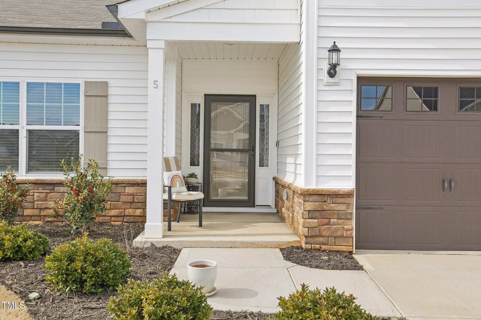 A small covered front porch with a chair, side table, and potted plants. White siding house with stone accents and a brown garage door. Number 5 visible on the wall next to the porch. Shrubs line the walkway.