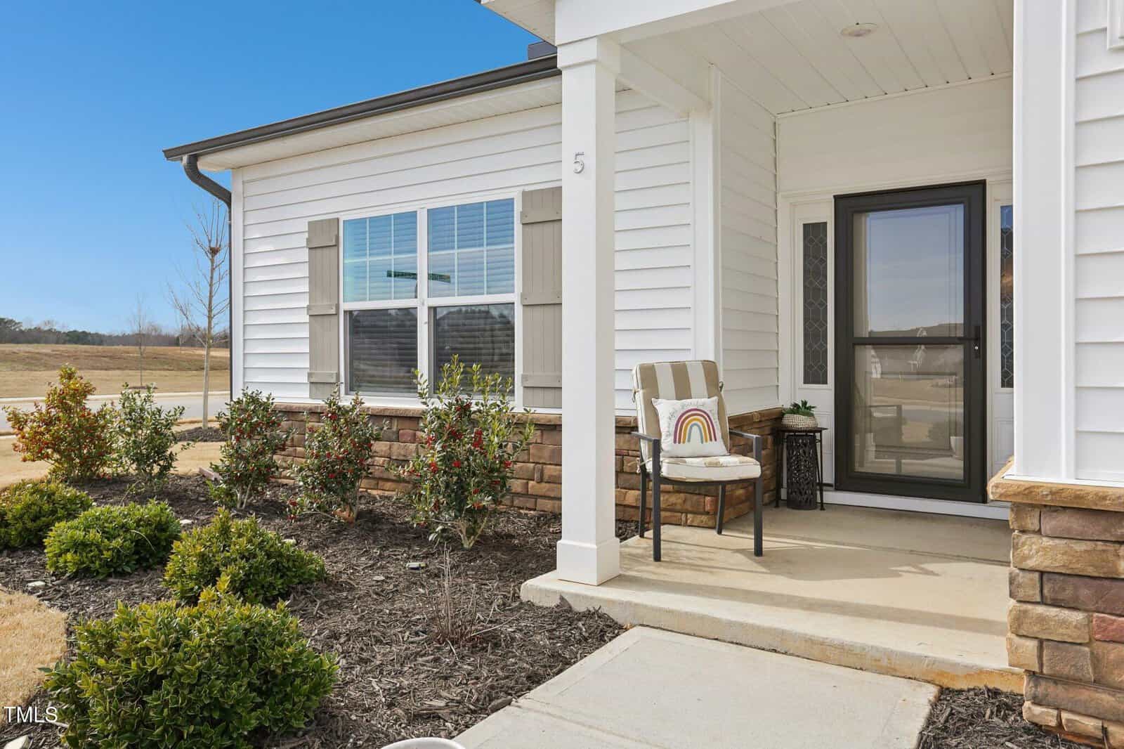 A small white house with stone accents, a covered porch, a glass front door, and a chair with striped cushion and rainbow pillow. Low shrubs are planted along the front walkway. Sky is clear and blue.