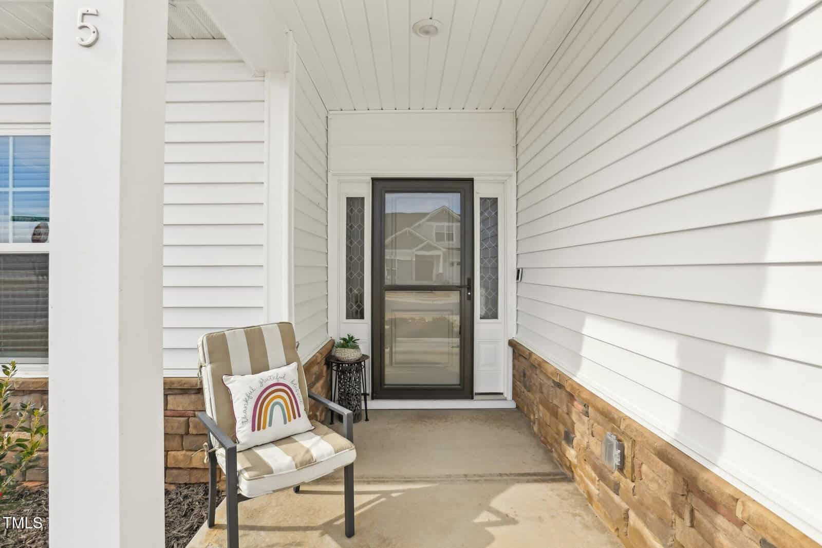 Covered front porch with white siding, glass storm door, small table with plant, and a striped chair holding a pillow featuring a rainbow design. House number 5 is visible on a column to the left. Brick trim along the bottom.