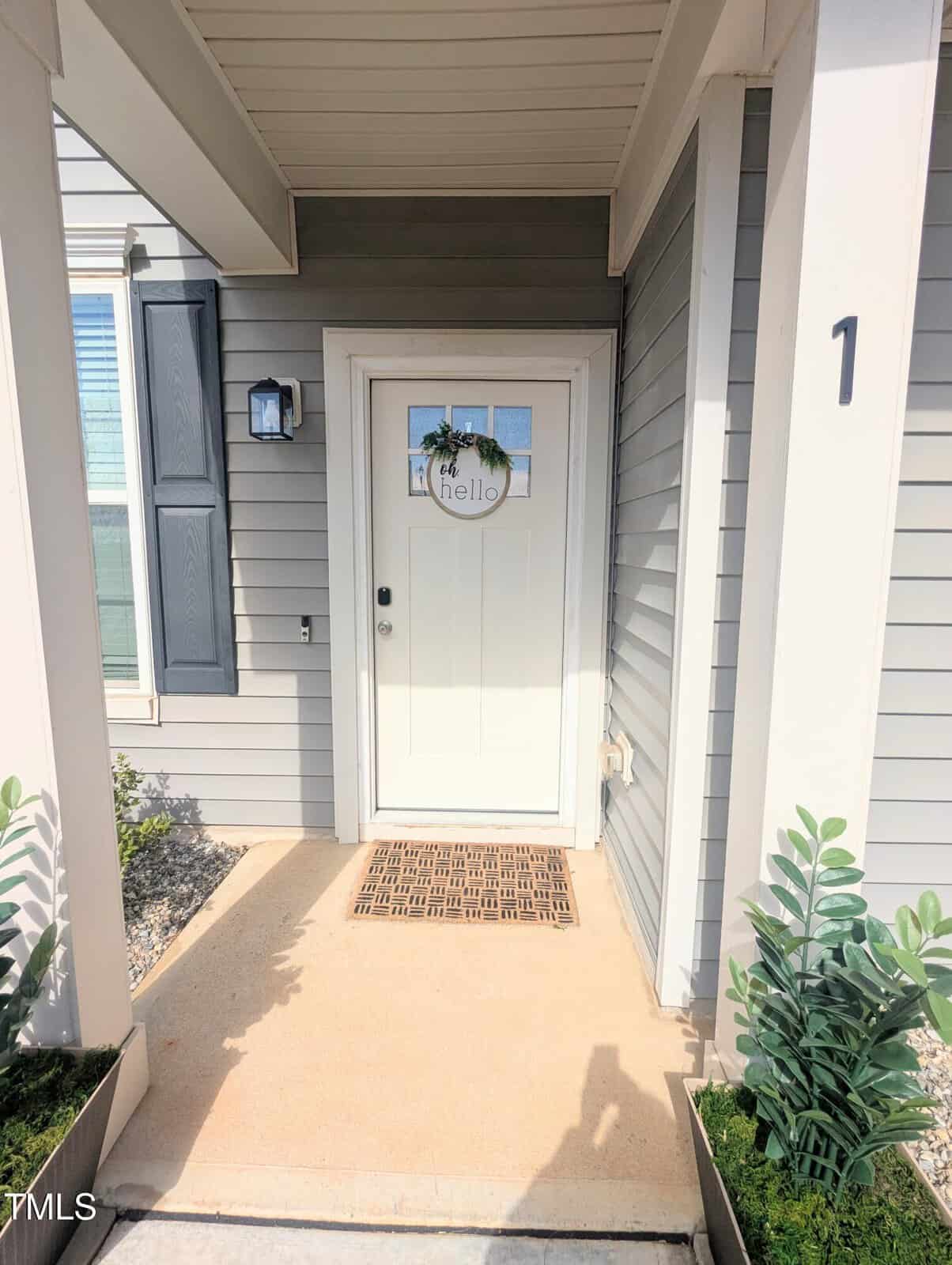 A white front door with a round "hello" sign and small wreath, framed by gray siding and white trim. There are two potted plants on either side of the entrance and a brown welcome mat on the porch.