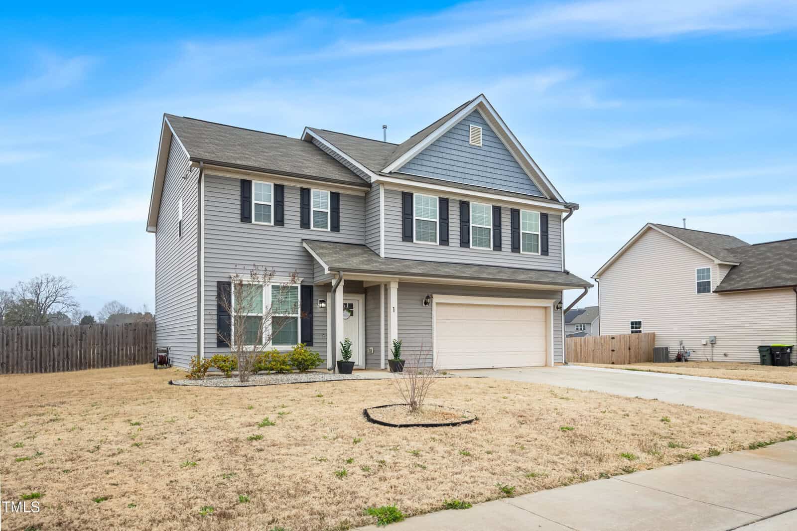 Two-story suburban house with gray siding, white trim, and a covered front porch, featuring a double garage, small front shrubs, and a large, dry lawn under a partly cloudy sky.