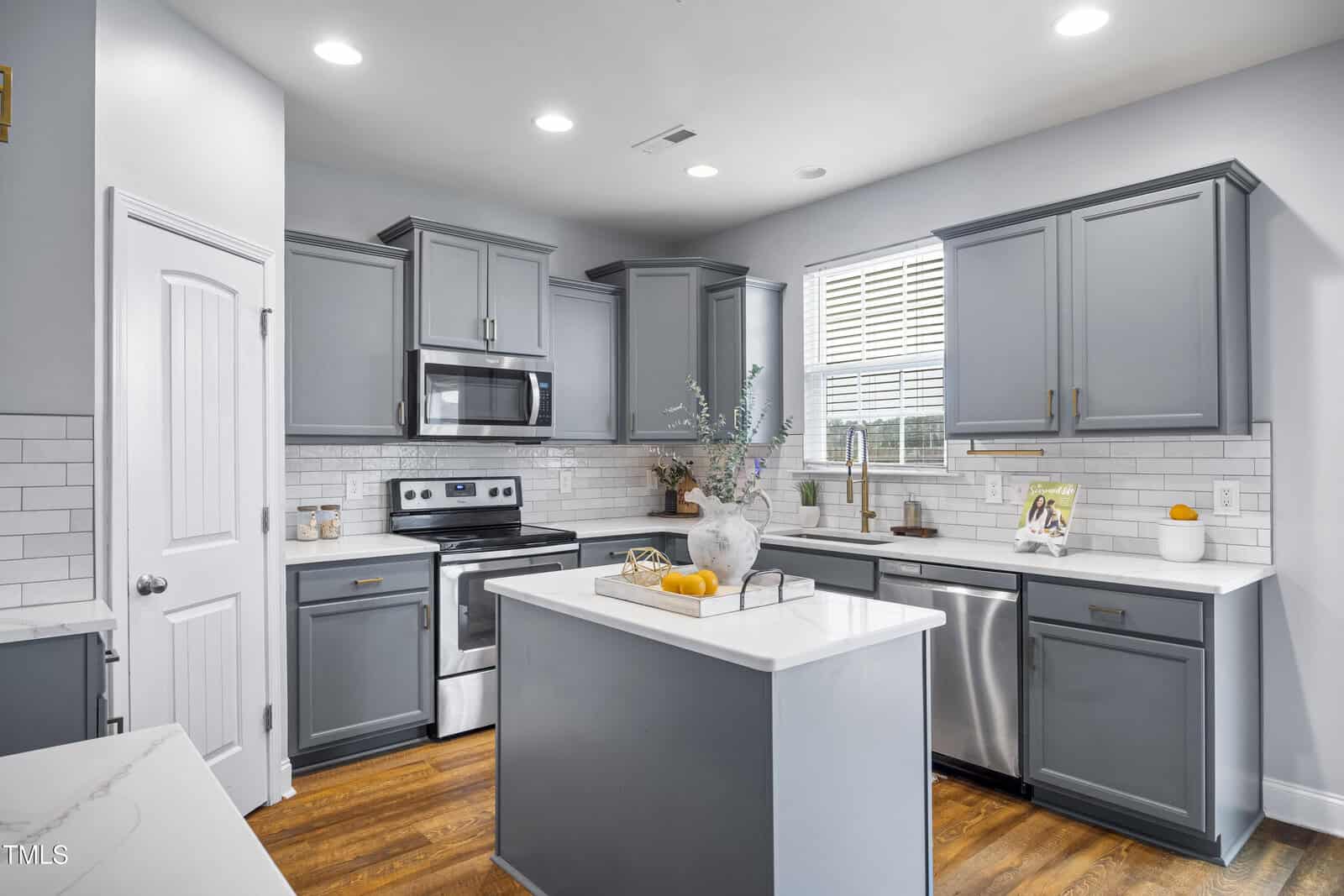 A modern kitchen with gray cabinets, stainless steel appliances, white subway tile backsplash, a center island with decor, and wood flooring. A window above the sink lets in natural light. Recessed lighting is on the ceiling.
