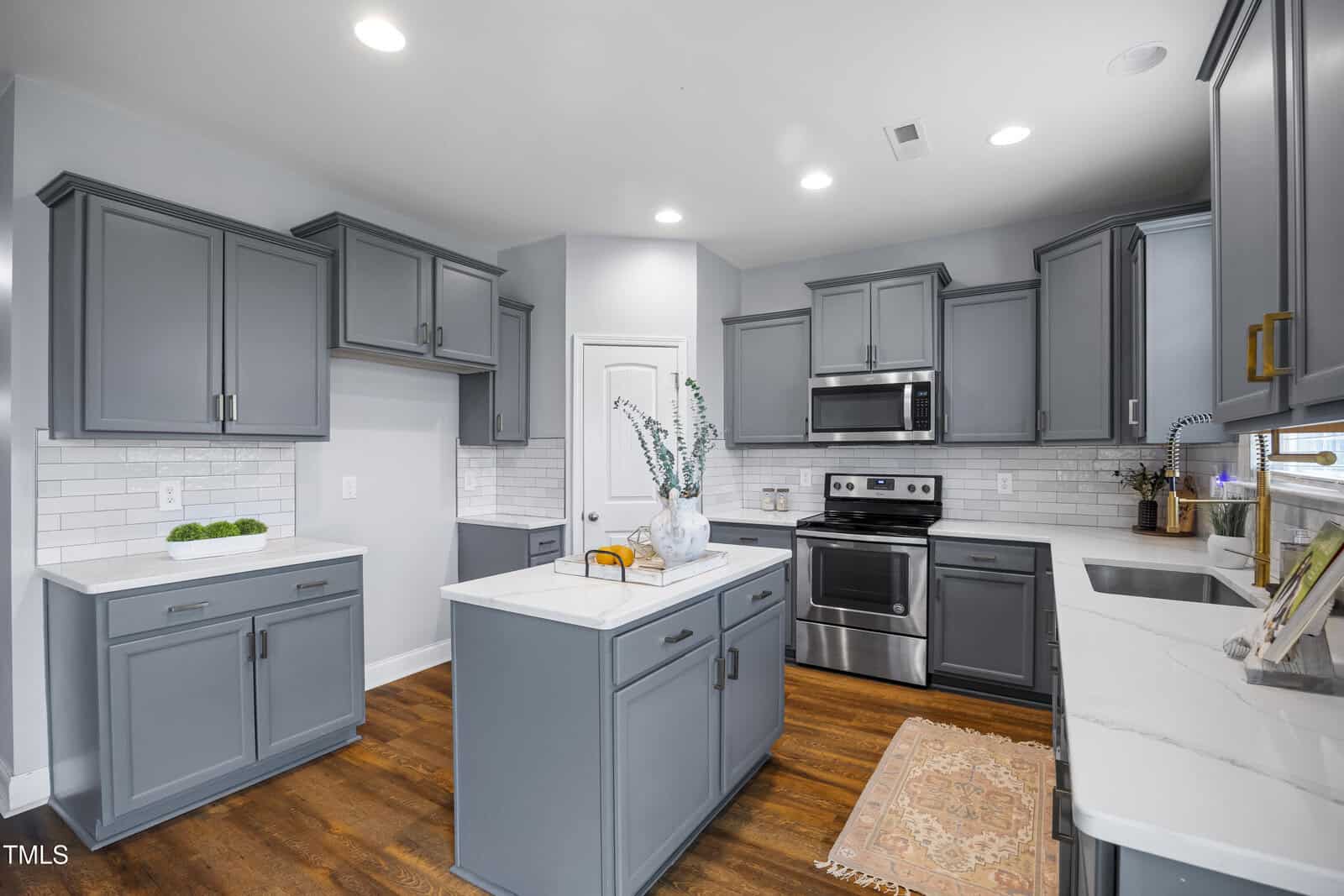 Modern kitchen with gray cabinets, white countertops, stainless steel appliances, a central island with a vase, wood floors, white subway tile backsplash, and a small rug near the sink. Bright lighting from ceiling fixtures.