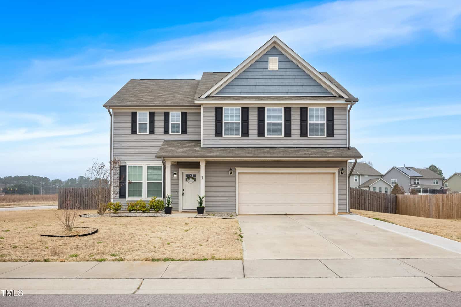 Two-story suburban house with gray siding, black shutters, and a double garage. The front yard has sparse grass, a few small bushes, and a concrete driveway under a clear blue sky.