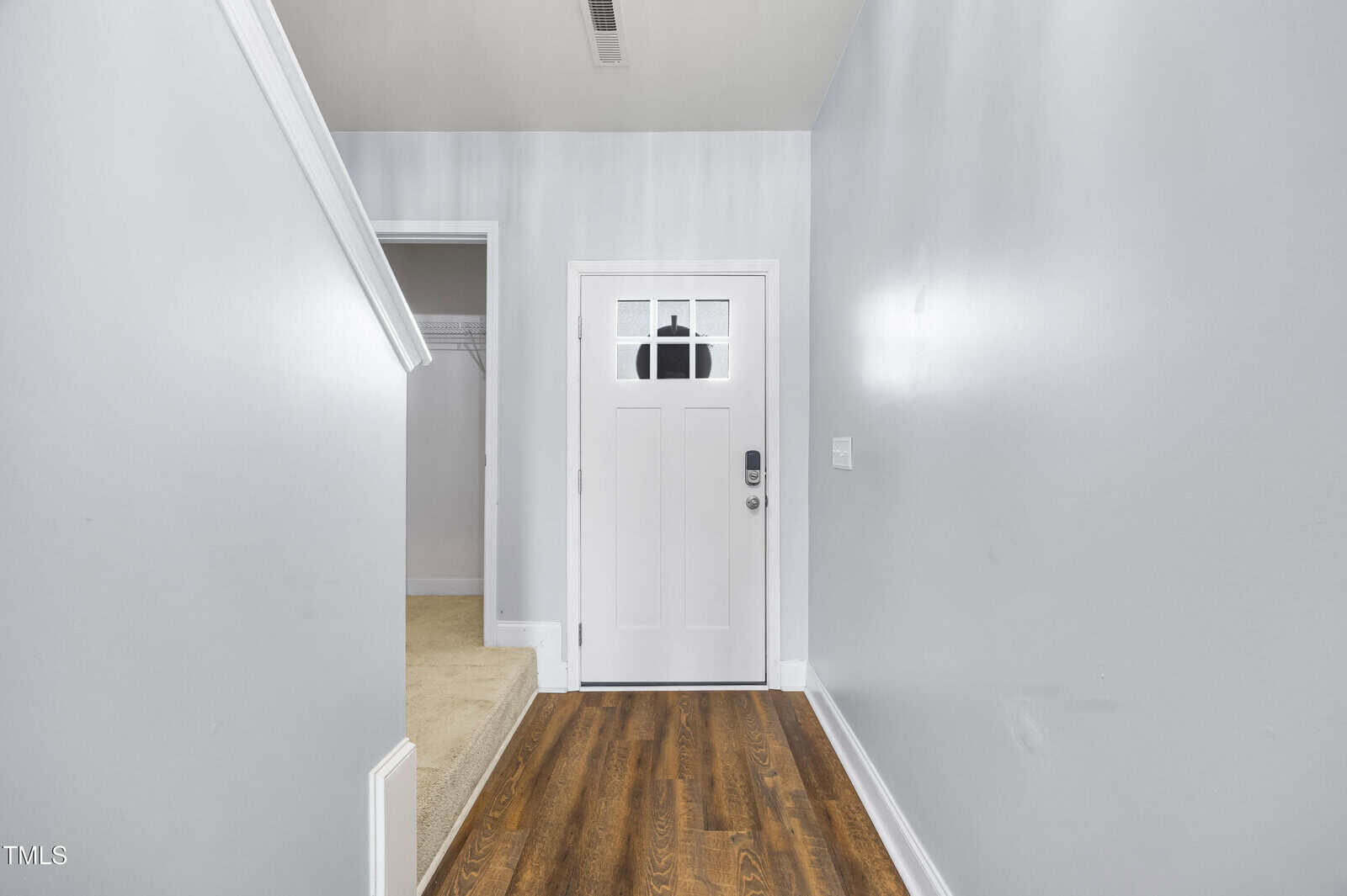 A front entryway with light gray walls, a white door with a window at the top, hardwood flooring, a carpeted closet to the left, and a staircase partially visible on the left side.