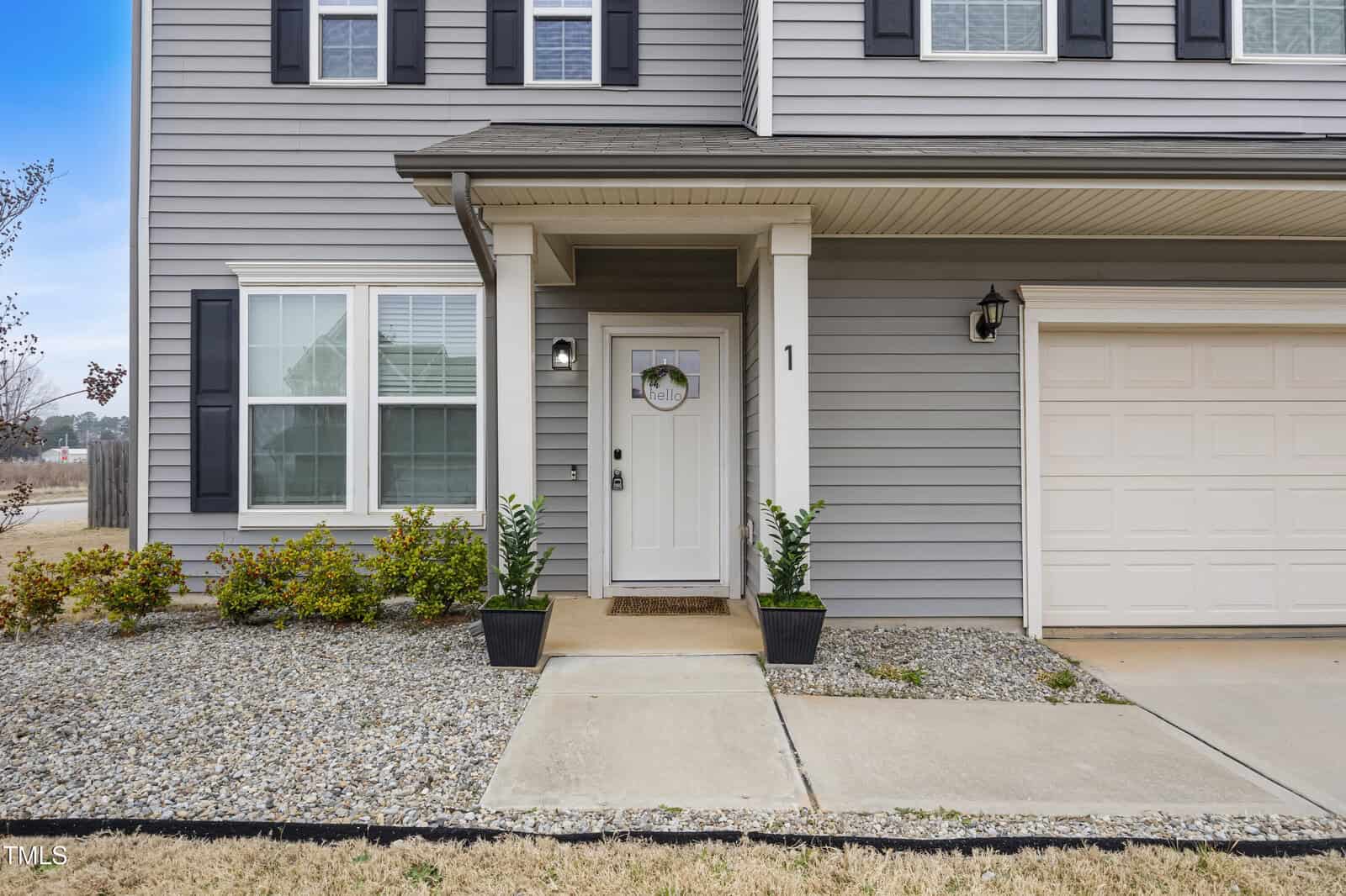 A gray two-story house with white trim, black shutters, and a white front door. Two potted plants sit by the entrance, and there is gravel landscaping with small shrubs near the door and window.