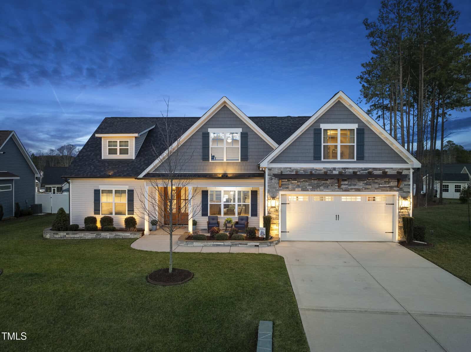 A two-story suburban house with gray siding, stone accents, and a double garage is shown at dusk with exterior lights on. The front yard is landscaped with grass, small shrubs, and a young tree.