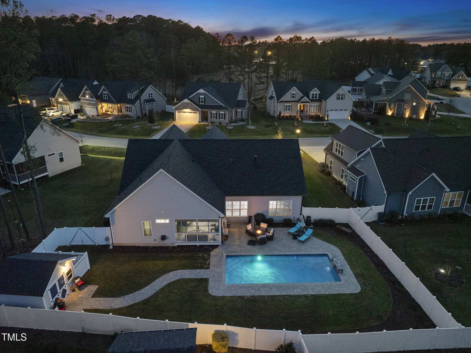 A fenced backyard at dusk features a lit in-ground swimming pool, a paved patio with outdoor seating around a fire pit, and a small pool house. Neighboring houses and trees are visible in the background.