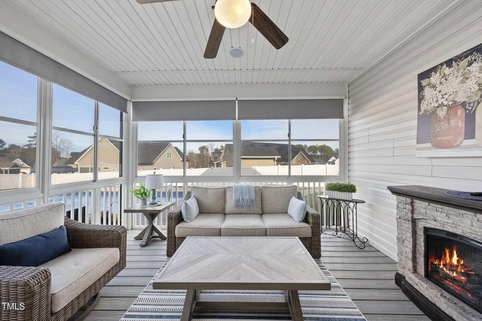 A sunroom features wicker furniture with gray cushions, a wooden coffee table, a striped rug, a ceiling fan, a fireplace with a floral painting above, and large windows overlooking neighboring houses.