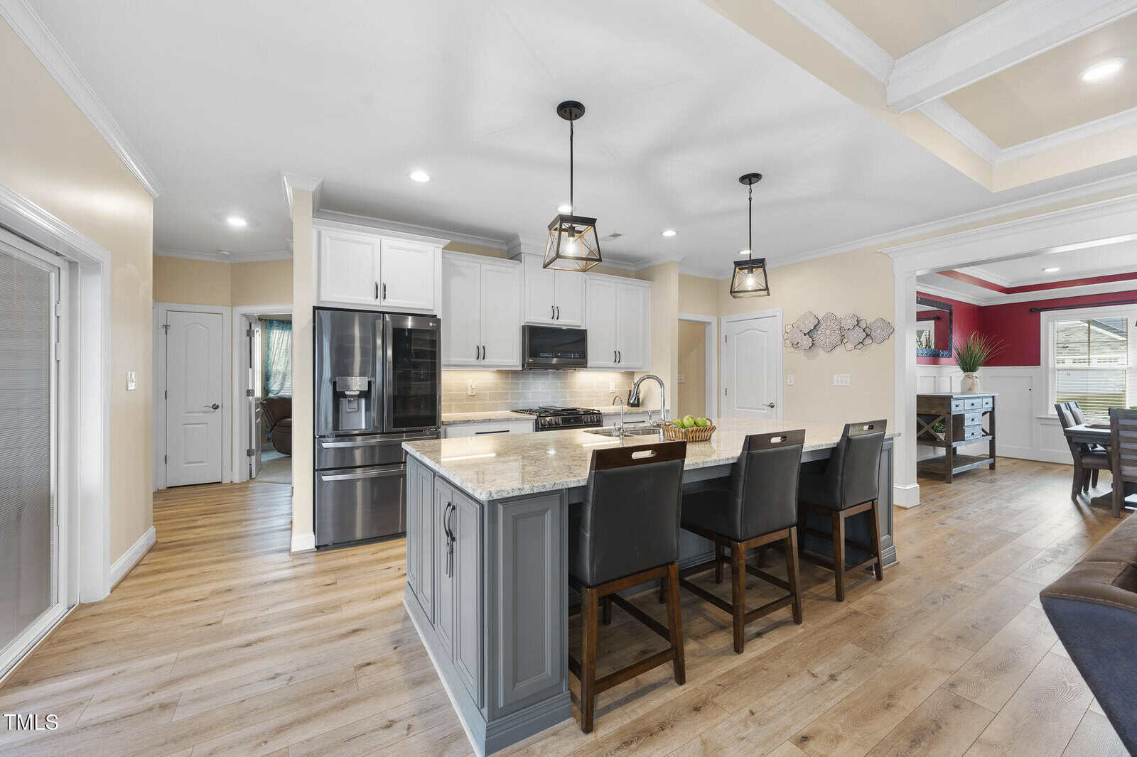 Modern kitchen with light wood floors, white cabinets, black appliances, a large island with granite countertop, four black chairs, pendant lights above, beige walls, and an open layout leading to a living area.