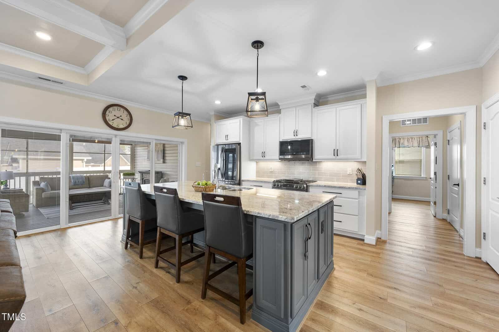 A modern kitchen with a large island, three dark chairs, white cabinets, stainless steel appliances, wood flooring, pendant lights, and sliding doors leading to a covered patio with seating.