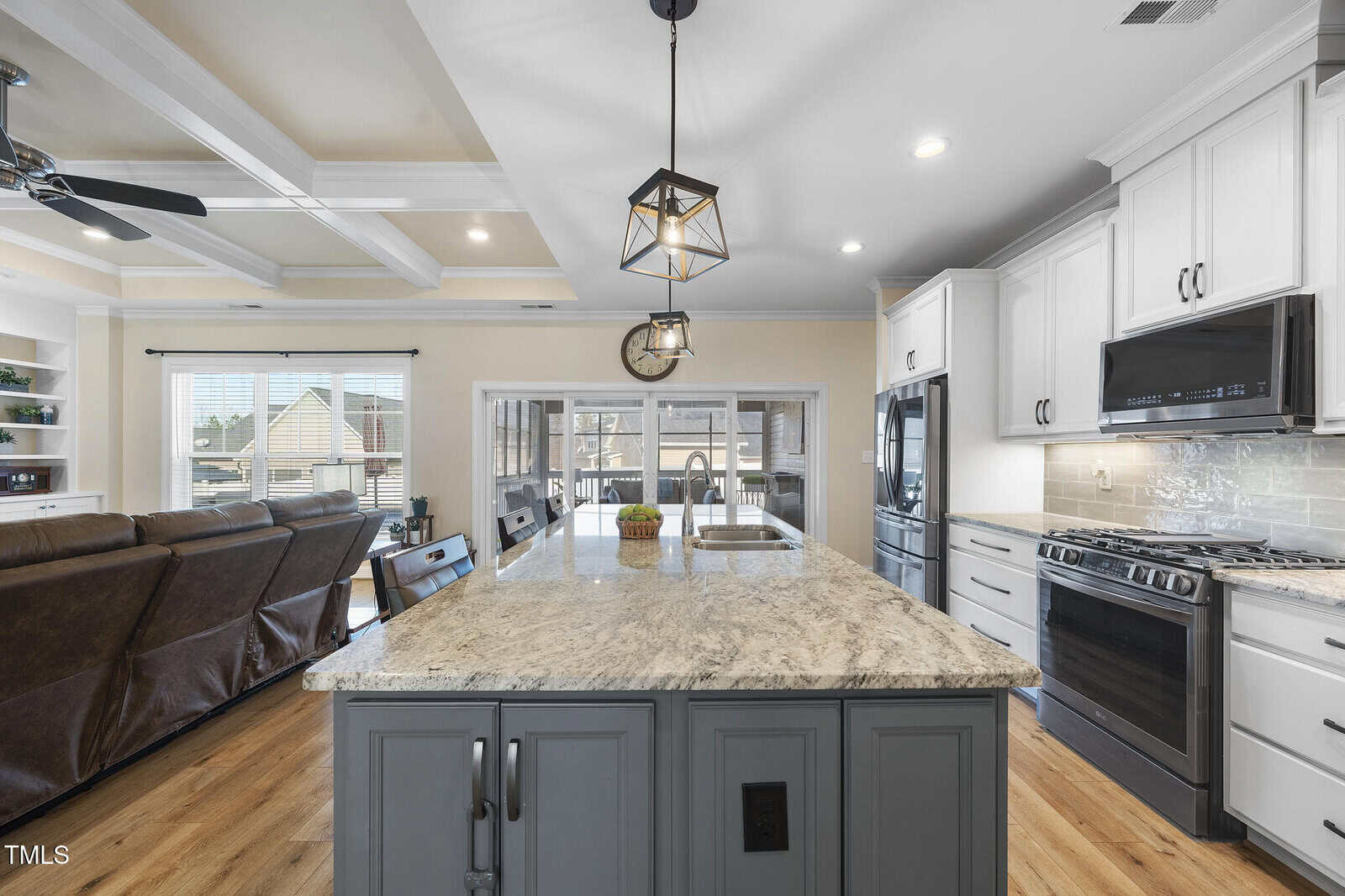 A modern kitchen with a granite island, white cabinets, stainless steel appliances, pendant lighting, and an open view to a living area with brown couches and large windows.