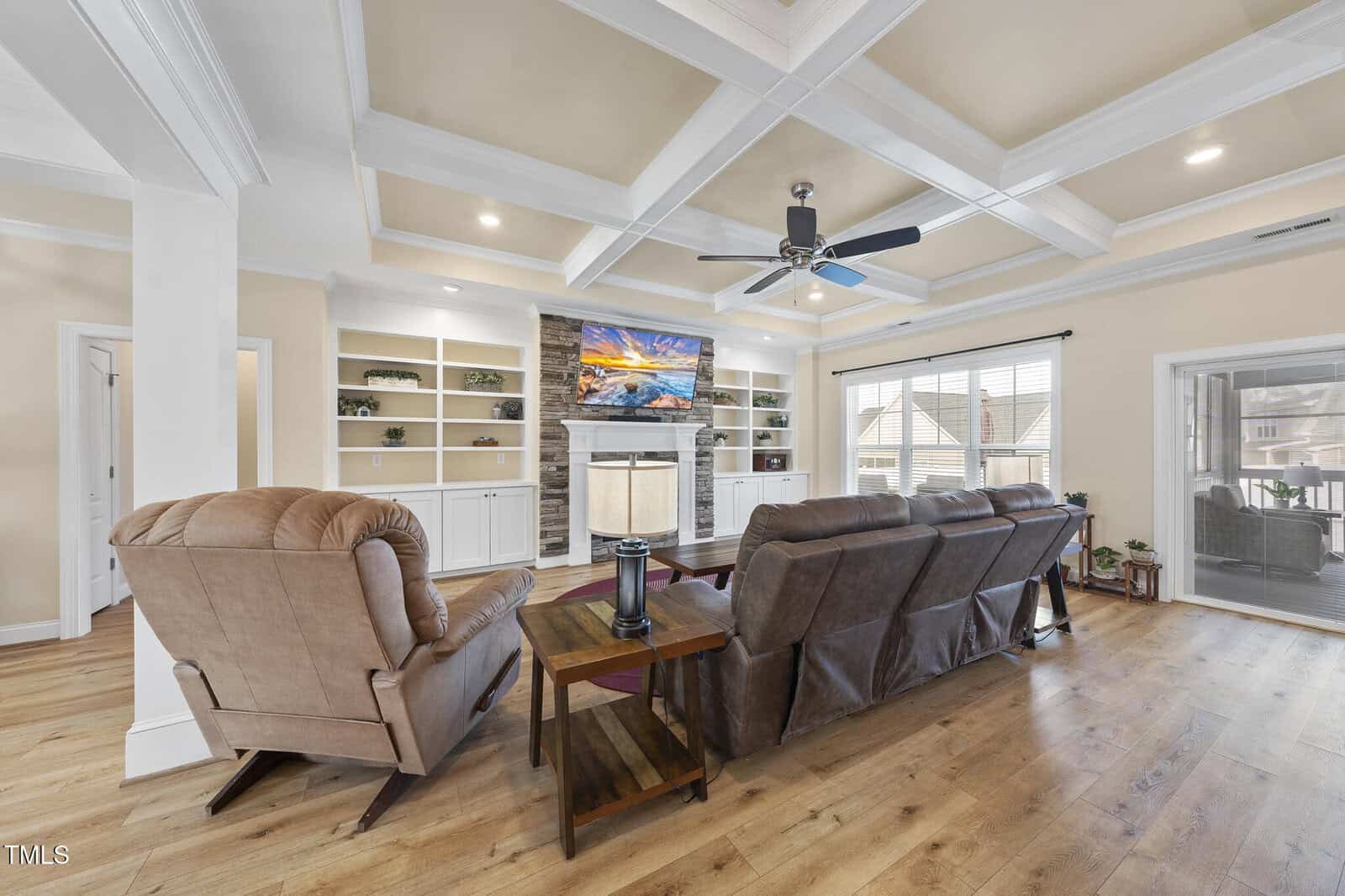 A living room with coffered ceilings, beige walls, wood flooring, two armchairs, a sofa, a ceiling fan, built-in shelves, a fireplace with a TV above it, and large windows letting in natural light.