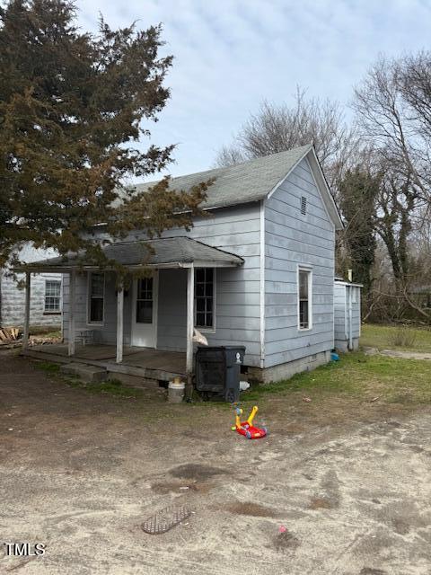 The weathered gray wooden house at 749 Southerland Street, Henderson has a porch, two small windows, and a sloped roof. A large tree towers beside it. Children's toys and a black trash bin clutter the entrance. The surrounding area is unkempt with patchy grass under an overcast sky.