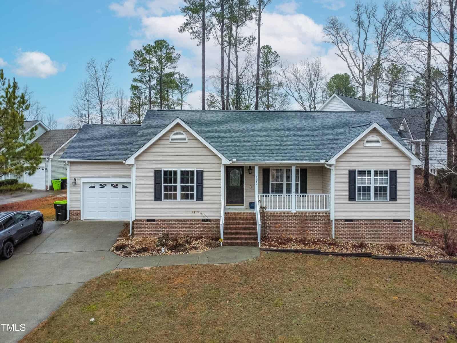 This charming single-story beige house at 2576 Mint Julep Drive features a dark roof and attached garage. A small front porch overlooks a simple lawn, with a black car parked in the driveway. Nestled in Creedmoor, trees frame the home against a cloudy sky.