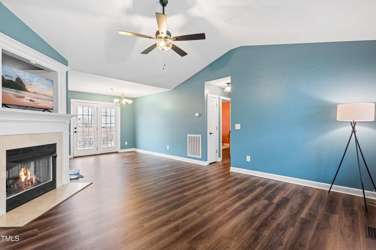 Living room with blue walls and wooden flooring. A lit fireplace is on the left, topped with a mounted TV. A ceiling fan with lights hangs centrally. There's a floor lamp on the right. French doors lead to the outside. An open doorway is visible.
