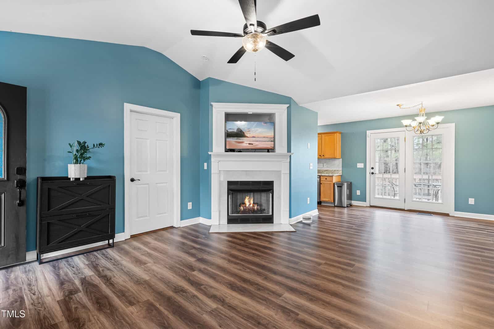 A spacious living room with blue walls and wood flooring. There's a ceiling fan, a fireplace with a TV above it, and a black console table beside a white door. The dining area and kitchen with wooden cabinets are visible in the background.