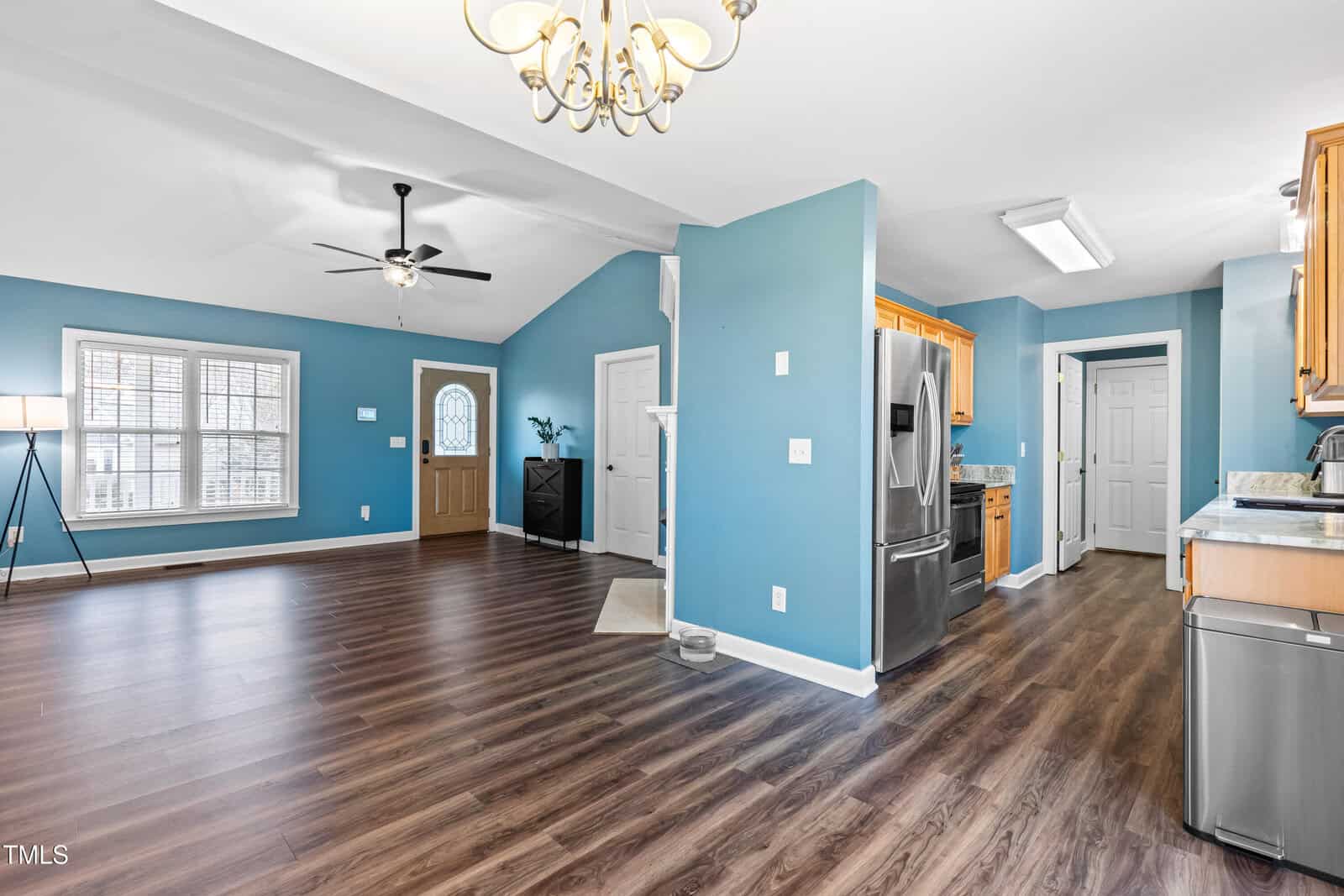 Spacious interior view of a home featuring an open living area with light blue walls and wood flooring. The kitchen is visible with stainless steel appliances and wooden cabinets. Ceiling fan, light fixture, and a window with blinds are present.